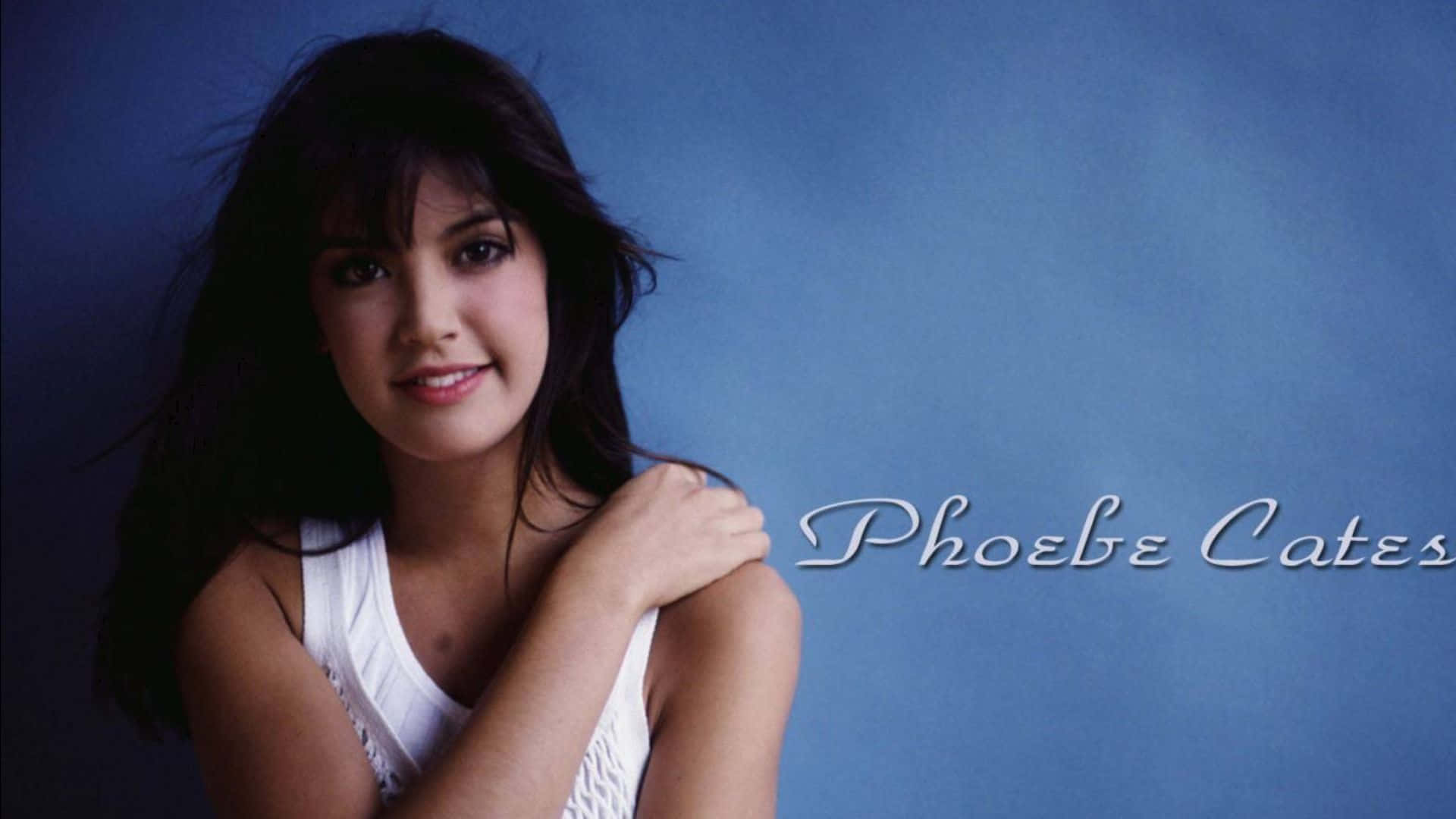 Phoebe Cates 桌布 壁紙 — 一位黑髮年輕女性在純藍色工作室背景前溫柔微笑，她的名字寫在旁邊。