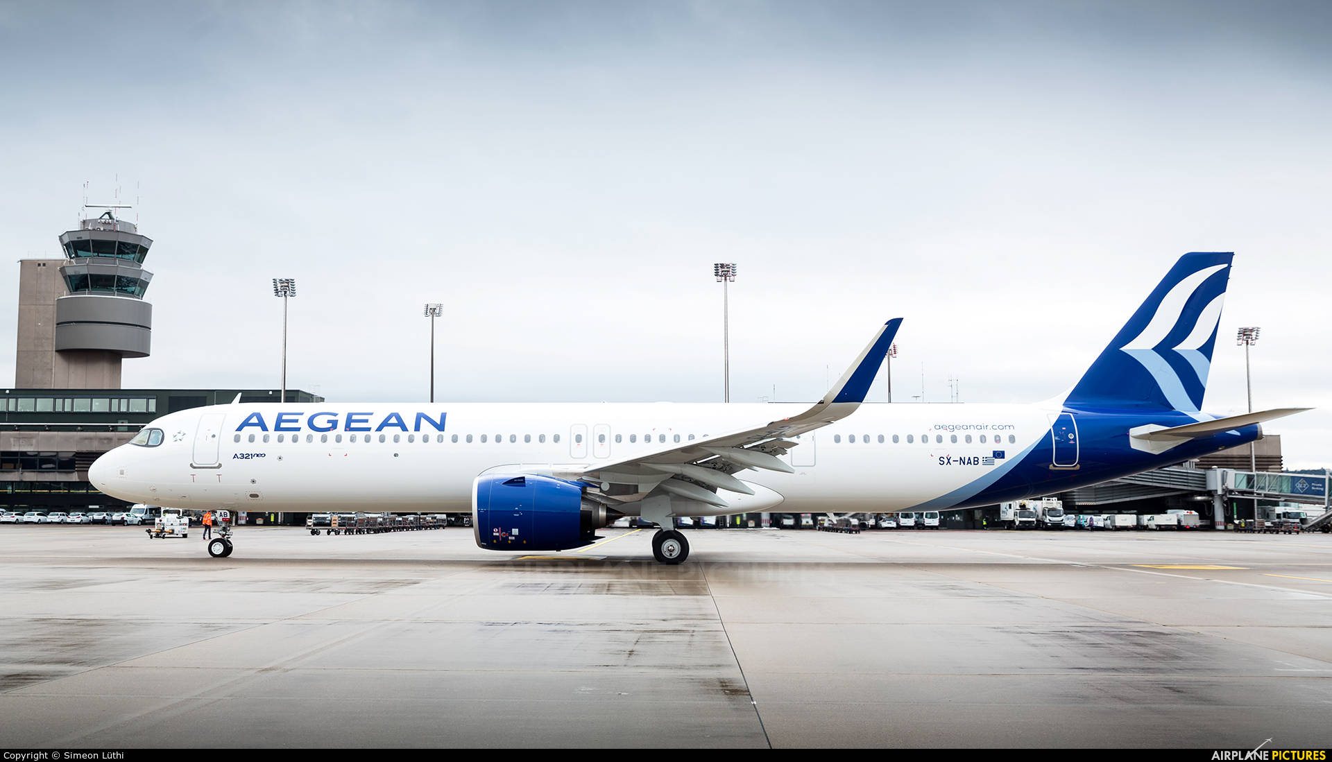 愛琴海航空A321neo飛行中與空管塔 壁紙 — 一架白色和藍色的愛琴海航空空中巴士A320停泊在濕滑的停機坪上，背景是機場控制塔，天空多雲。