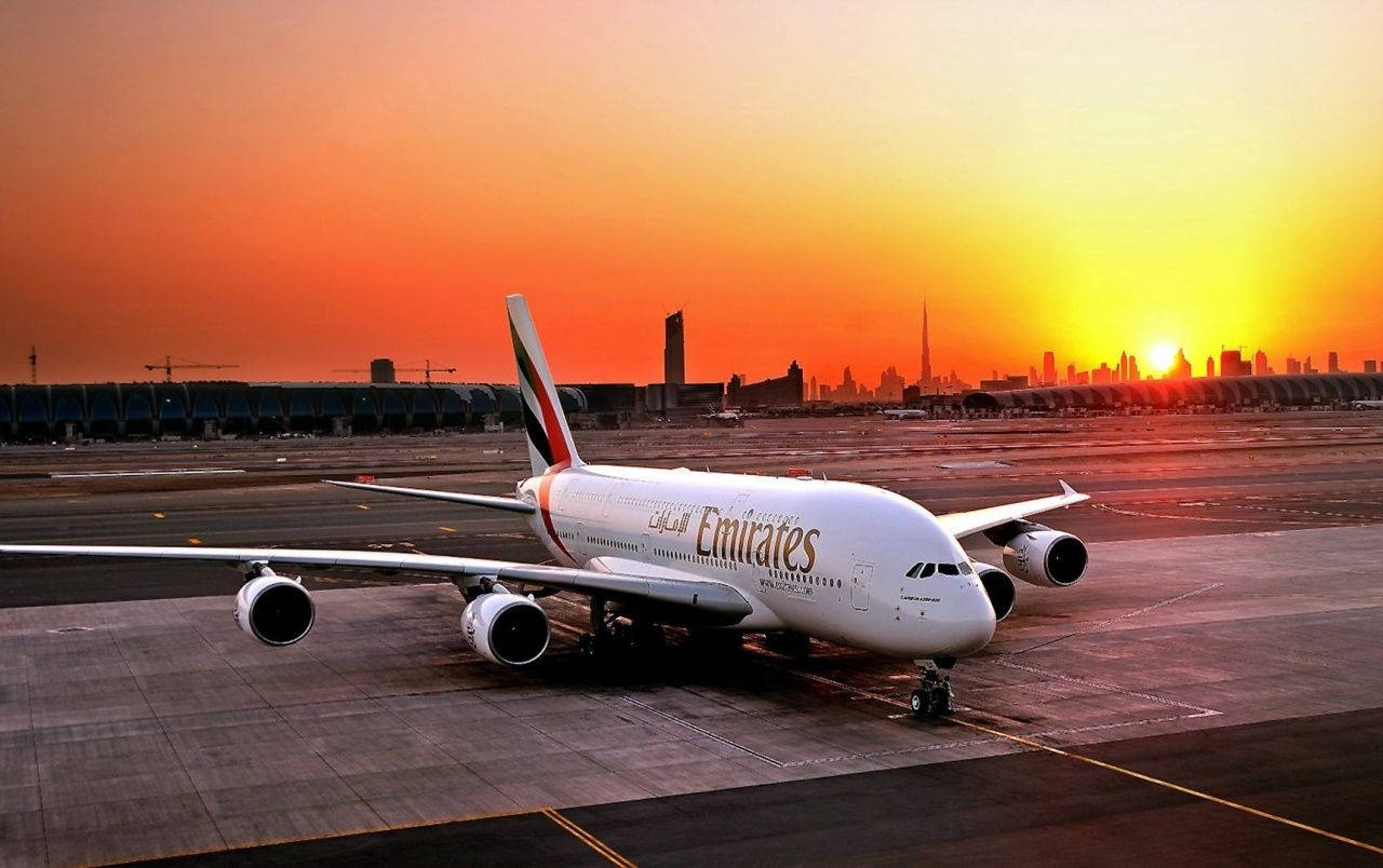 阿聯酋航空A380與壯麗日落 壁紙 — 一架阿聯酋航空的空中巴士A380停在機場停機坪上，夕陽在現代城市天際線後落下，散發出溫暖的橙色和黃色光芒。