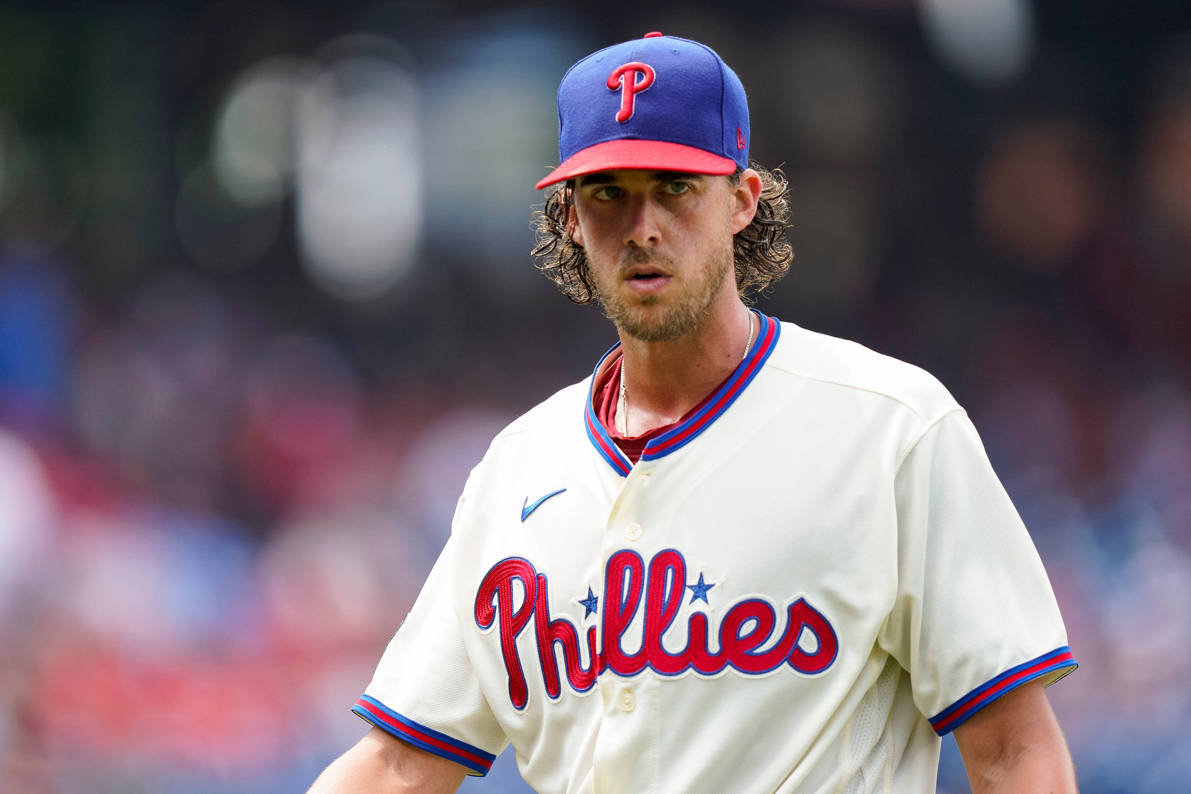 Aaron Nola 凝視 壁紙 — 一位專注的費城費城人棒球運動員，身穿白色配紅藍邊的球衣，在陽光明媚的球場上比賽。
