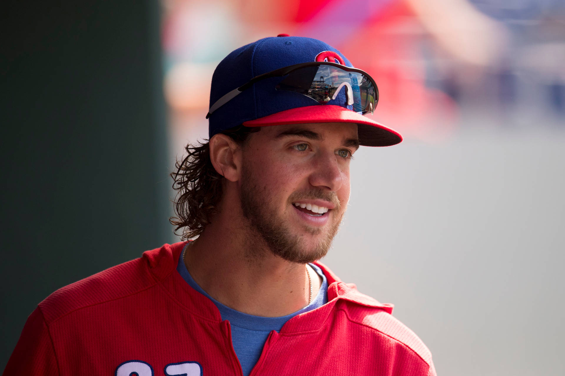 Aaron Nola 微笑 壁紙 — 一位微笑的棒球運動員，身穿紅色球衣和藍色帽子，帽子上架著太陽眼鏡，正望向一旁。