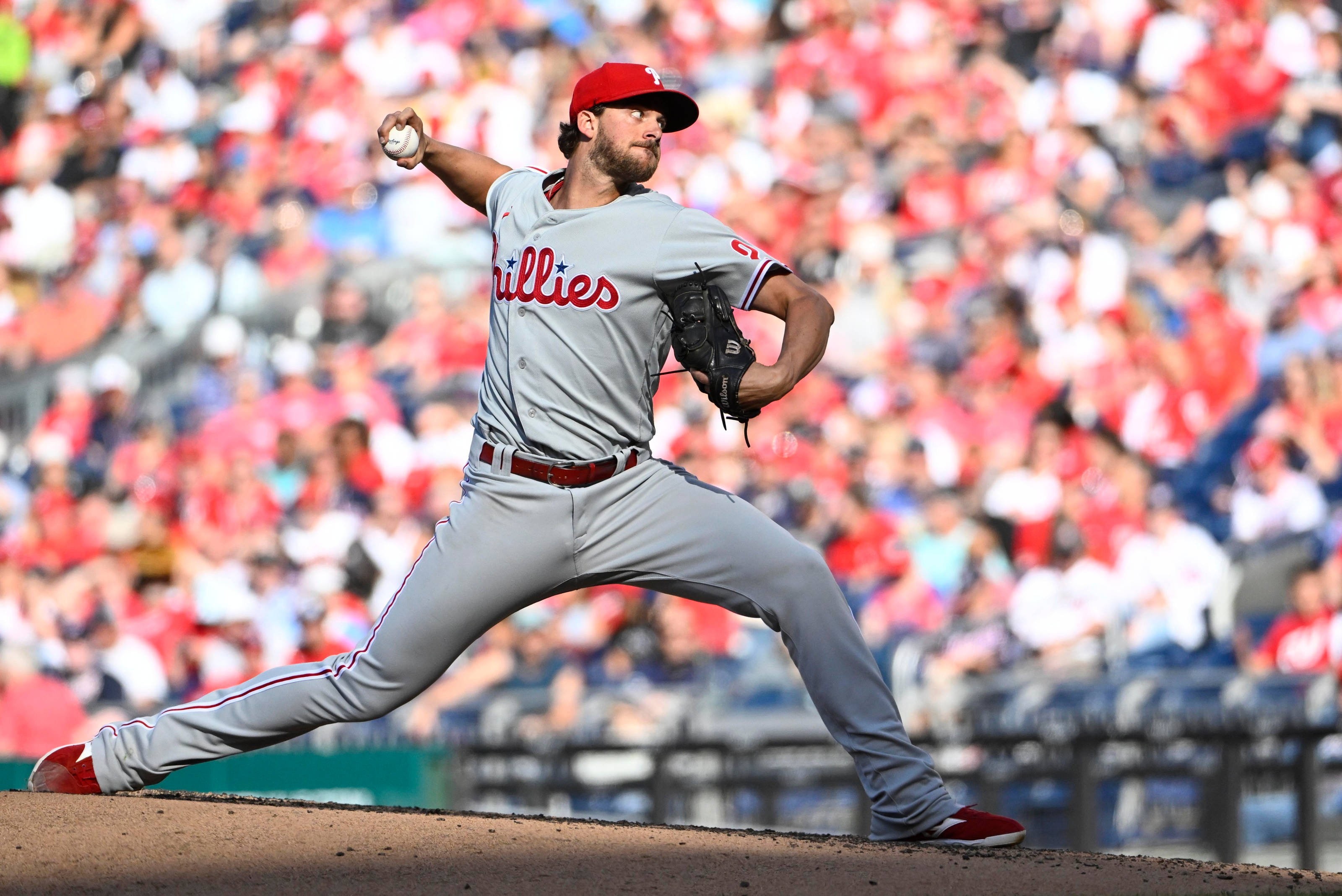 Aaron Nola 投球 壁紙 — 一名身穿灰色費城人隊球衣的棒球投手，在模糊的紅白球迷人群中，專注地投球。