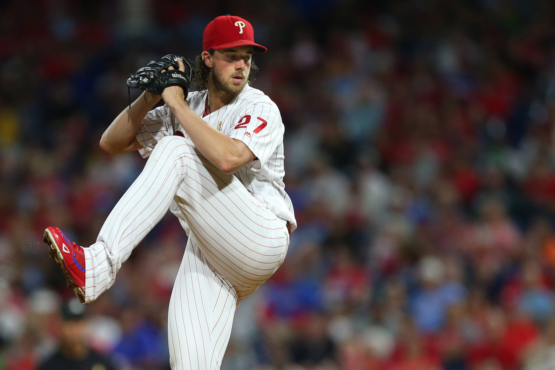 Aaron Nola 瘋狂蜷縮 壁紙 — 一位身穿白色細條紋制服、戴紅色帽子、背號27的棒球投手，正處於投球動作中，一條腿高高抬起，背景是模糊的體育場人群。