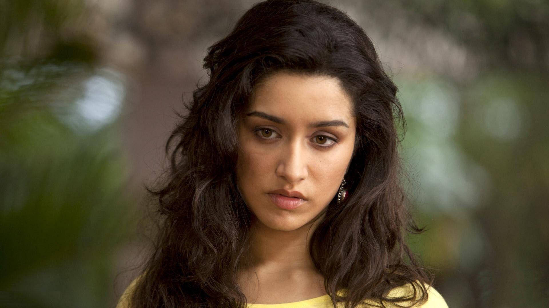 Aashiqui 2 阿蘿希卷髮特寫 壁紙 — 一位擁有深色波浪頭髮和黃色上衣的女性，在模糊的綠色樹葉背景下專注地凝視。