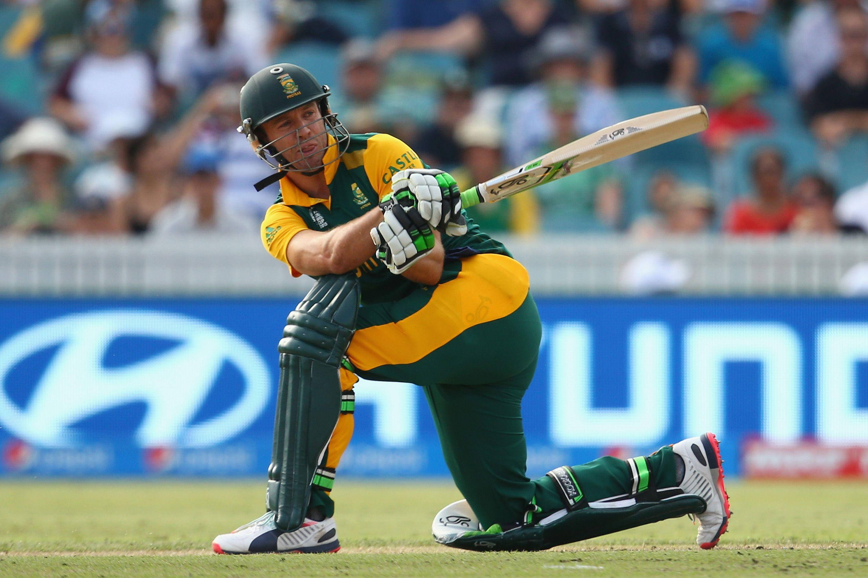 AB de Villiers 強力揮棒 壁紙 — 一名身穿綠色和黃色球衣的板球擊球手在陽光明媚的球場上揮動球棒，背景有模糊的觀眾。