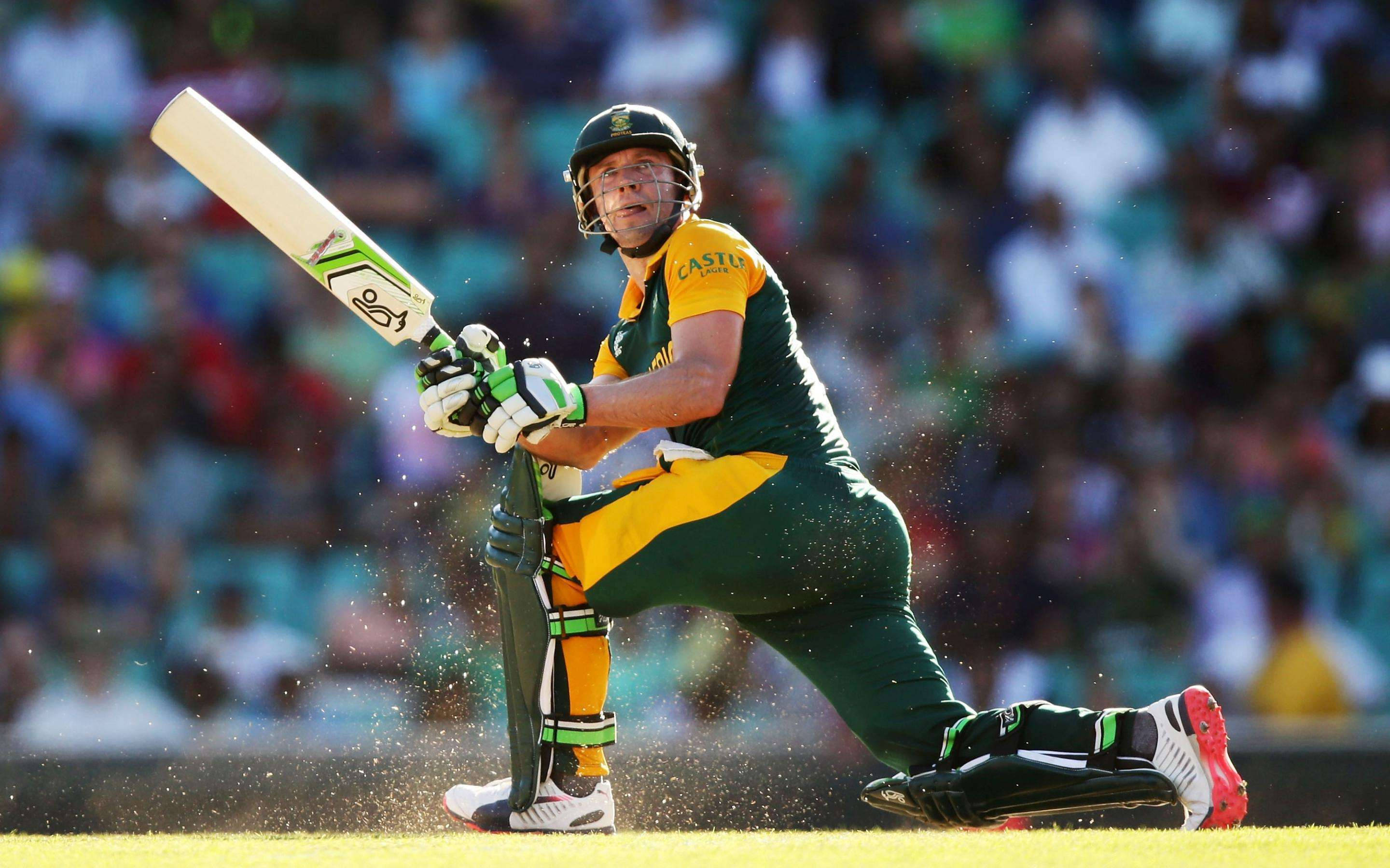 AB de Villiers 汗流浹背蹲伏 壁紙 — 一名身穿綠色和黃色球衣的板球運動員在陽光明媚的球場上向前猛衝，球棒和球在運動中揮動。