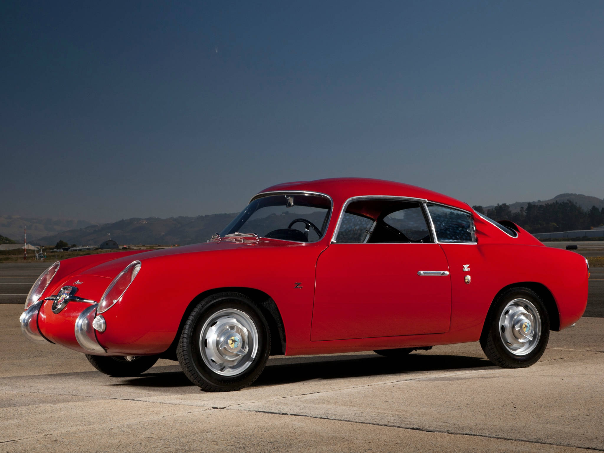 菲亞特Abarth 750 Zagato 壁紙 — 一輛鮮紅色復古跑車，帶有鍍鉻裝飾，停放在混凝土停車場上，背景是晴朗的藍天和遠山。