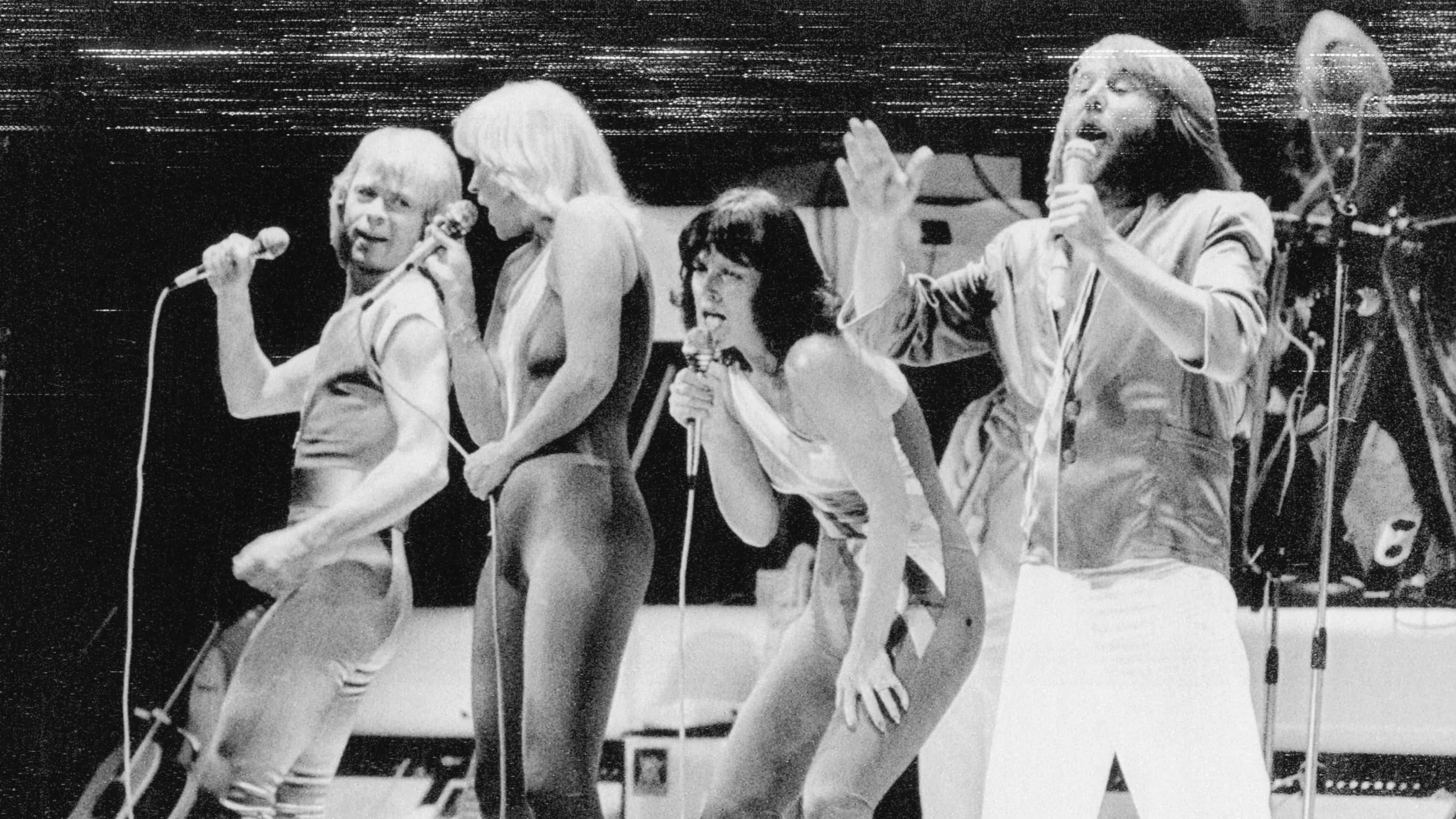 傳奇迪斯可樂團「ABBA」 壁紙 — 四位穿著緊身連體衣的表演者在明亮的燈光下，在充滿活力的現場音樂會中於舞台上熱情歌唱。