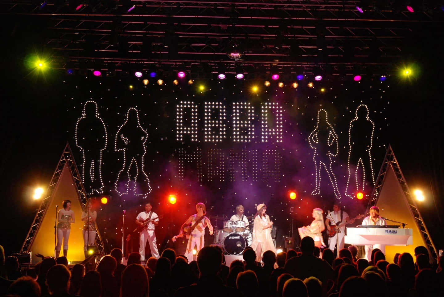 經典音樂傳奇ABBA 壁紙 — 一支ABBA致敬樂隊在色彩繽紛的舞台燈光下表演，背景黑暗，帶有剪影和LED文字。