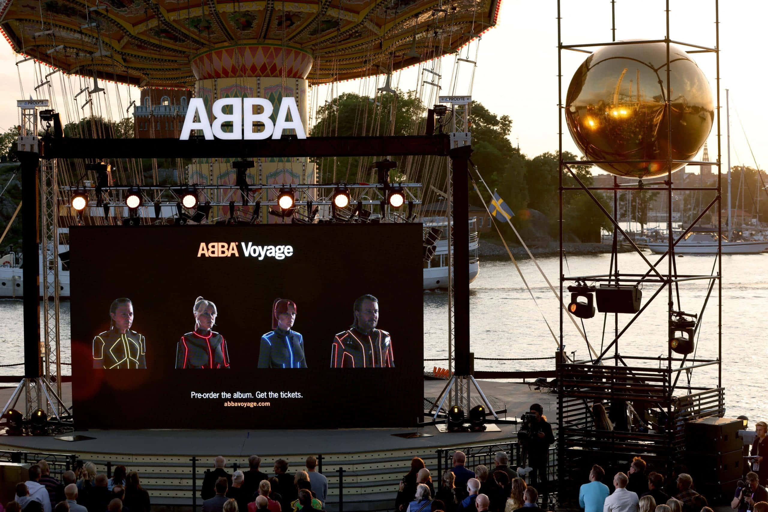 音樂傳奇ABBA與他們的經典名曲 壁紙 — 大屏幕顯示ABBA Voyage，四位表演者穿著未來主義服裝，在日落時分的海濱舞台上演出。
