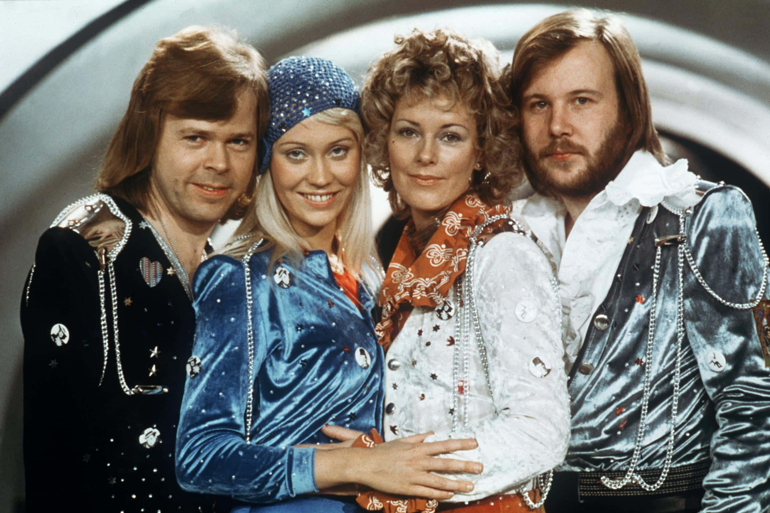 傳奇樂團ABBA 壁紙 — 四位1970年代華麗的音樂家穿著閃亮的天鵝絨外套，在彎曲的金屬背景前自信地擺姿勢。