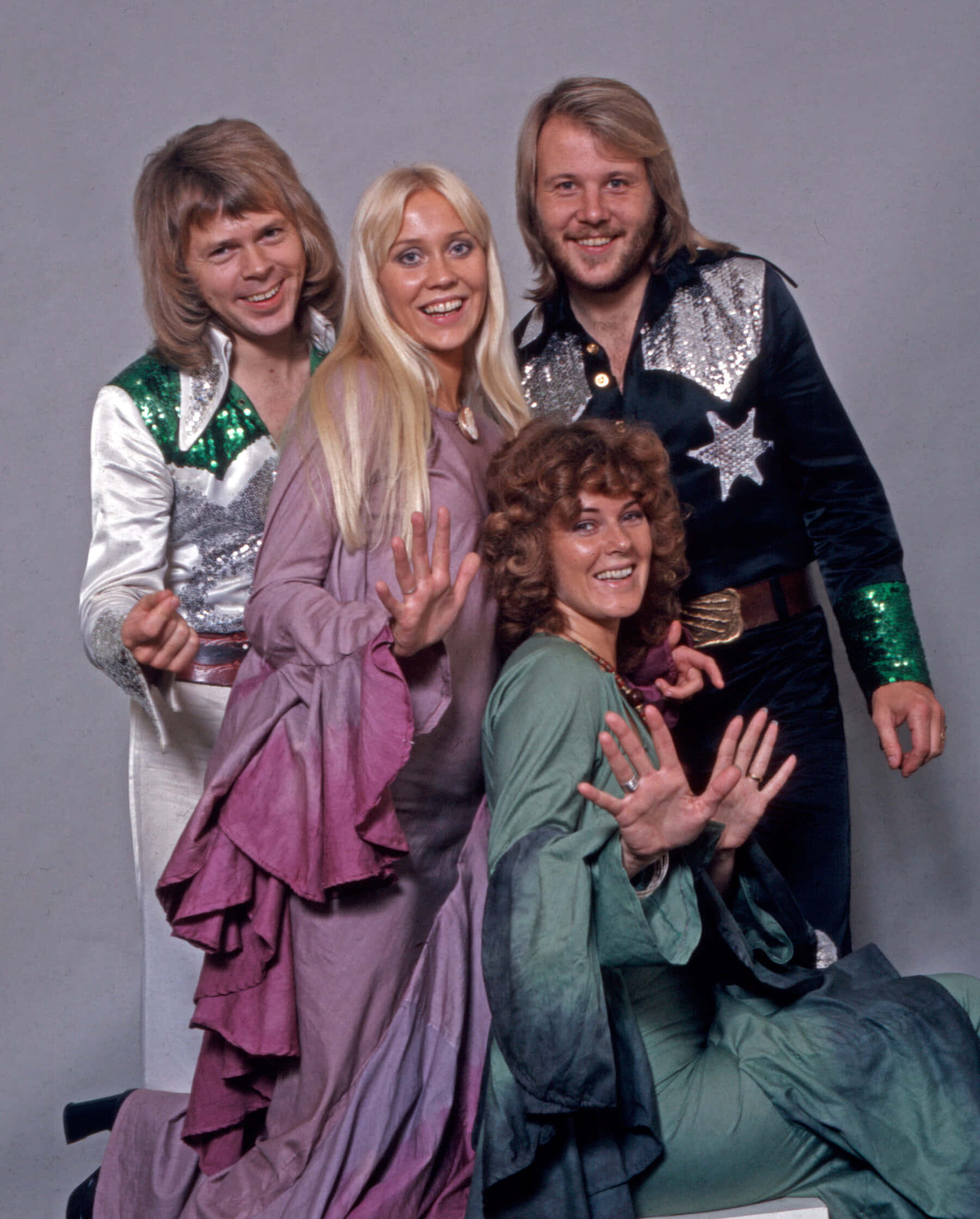 傳奇流行樂團ABBA 壁紙 — 四位微笑的1970年代音樂家穿著閃閃發光的亮片服裝，在樸素的灰色工作室背景前愉快地擺姿勢。