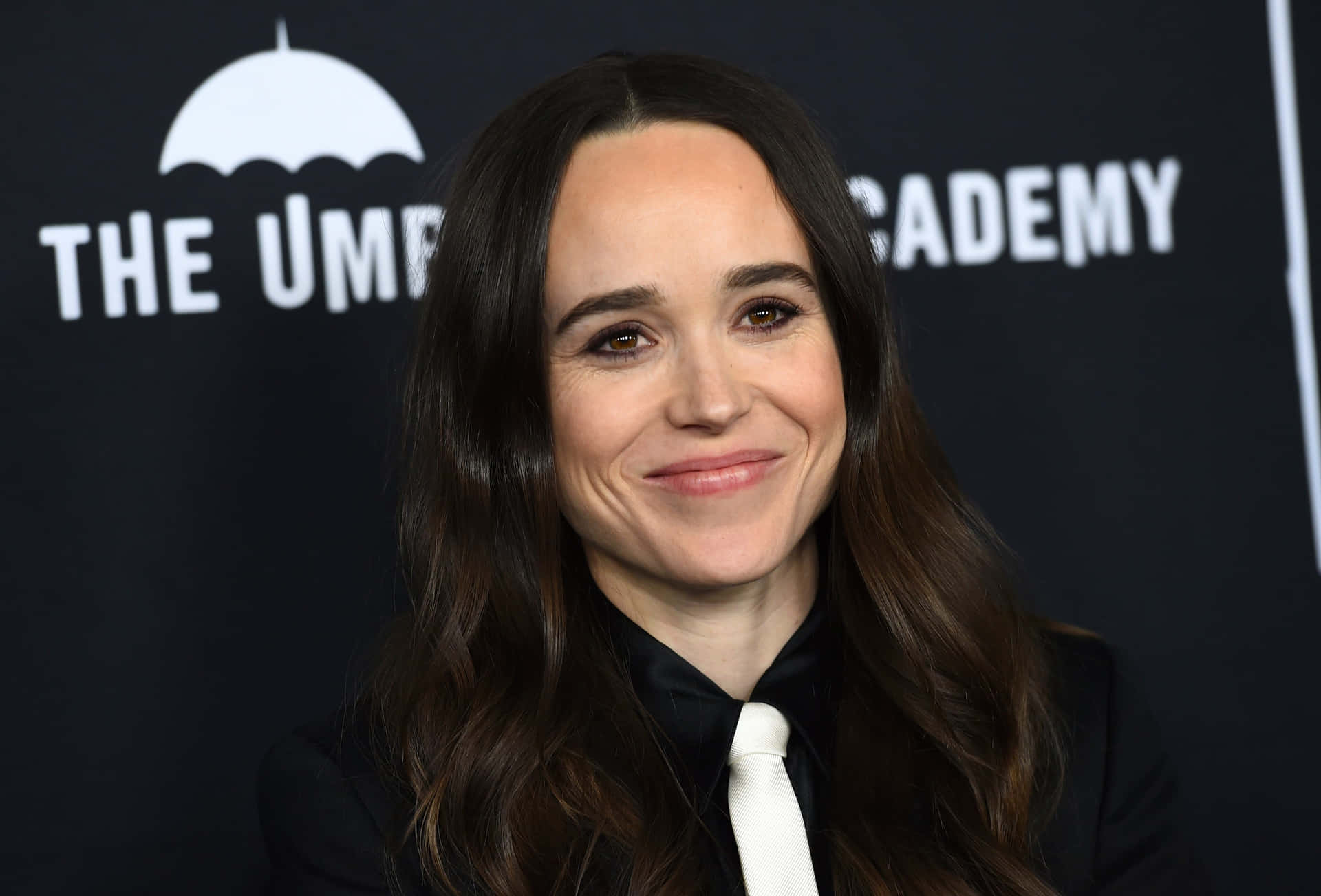  壁紙 — 女演員艾倫·佩姬穿著黑色西裝和白色領帶，在帶有「The Umbrella Academy」標誌的深色背景下，對著鏡頭溫暖地微笑。