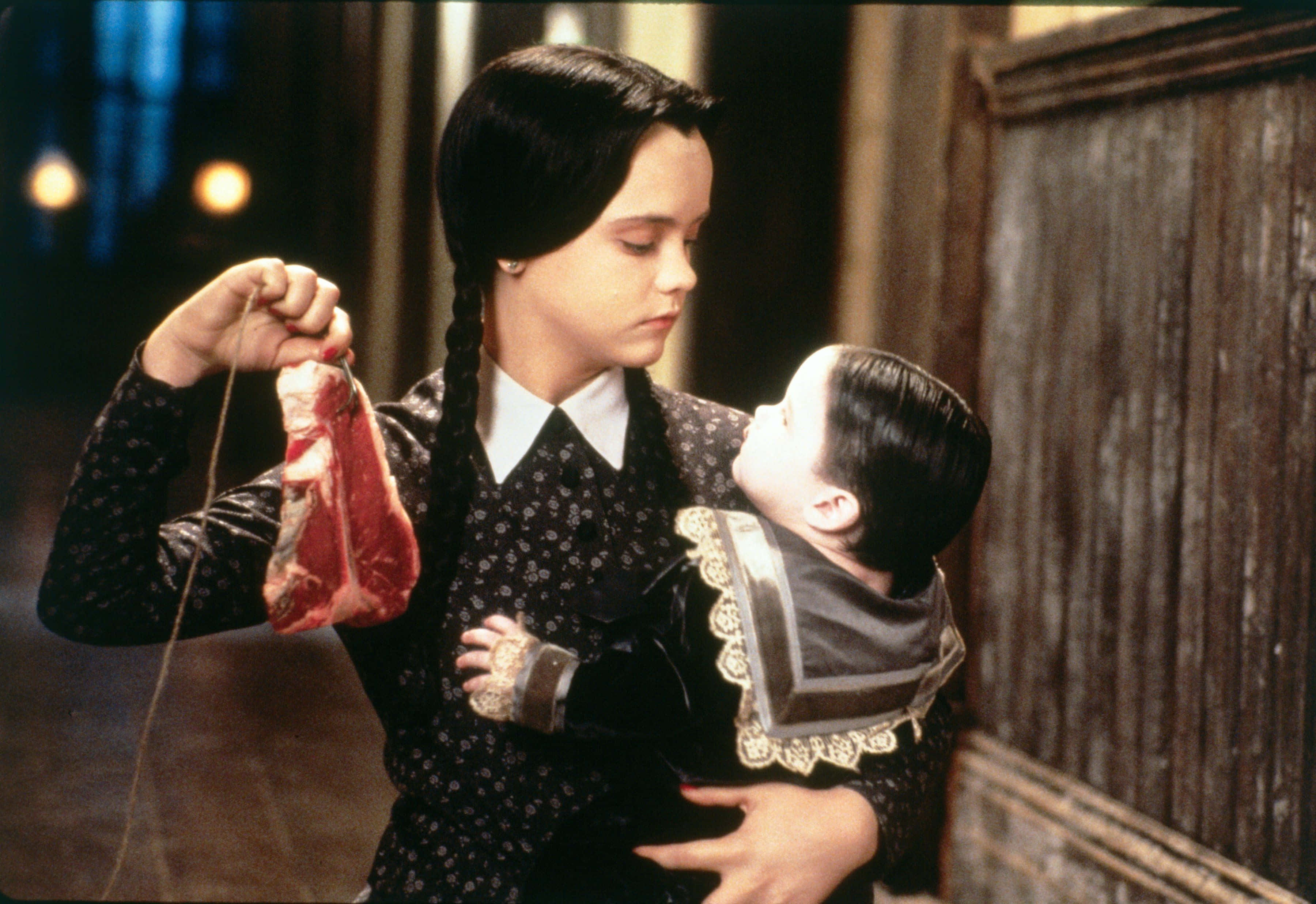 《Addams Family》——加入這古怪又迷人的世界 壁紙 — 一位身穿哥德式服裝的年輕女孩，在光線昏暗的木牆房間裡，抱著一個嬰兒，同時拿著一包生肉。