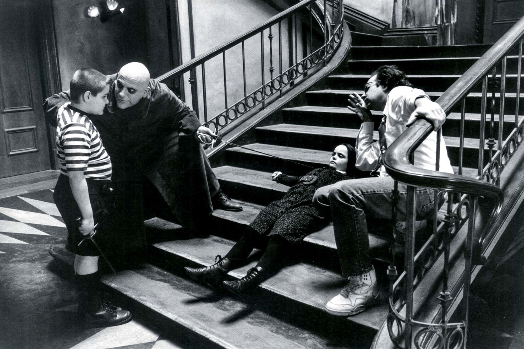 《Addams Family》聚集在他們詭異宅邸的經典客廳 壁紙 — 一個黑白場景，四個角色在哥德式豪宅的華麗樓梯上，帶有戲劇性的燈光。