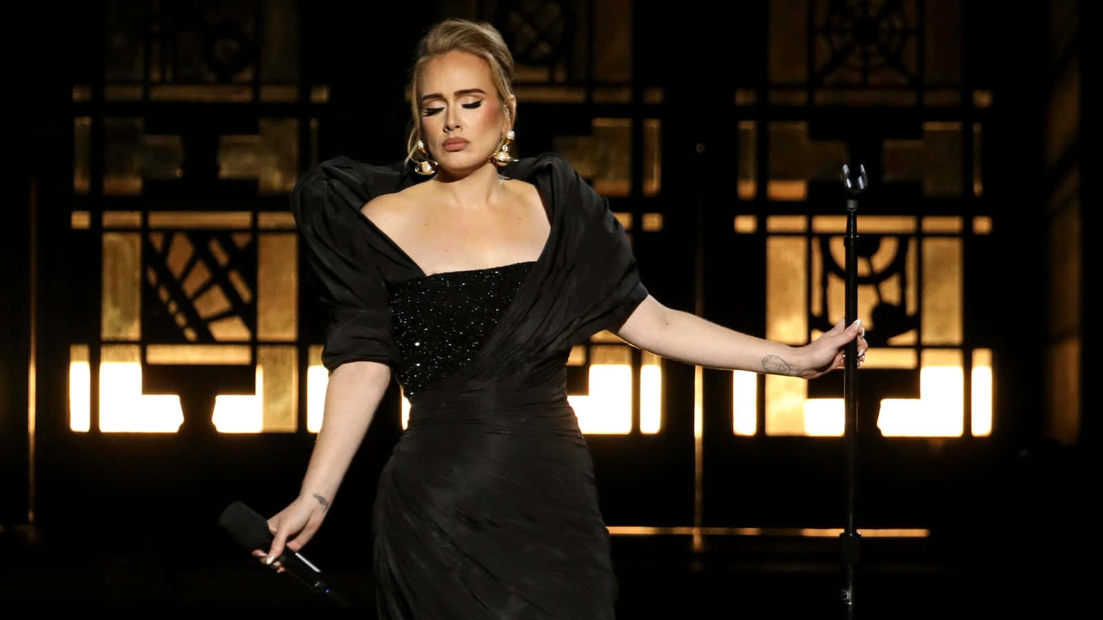 Adele 身穿黑色連衣裙在舞台上唱歌，身後有金色燈光。