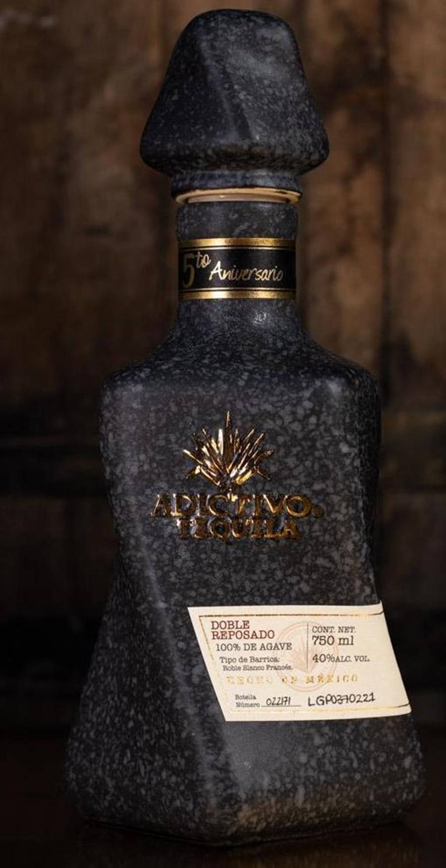 Adictivo 陳年龍舌蘭酒 黑色版 壁紙 — 一個深色斑點龍舌蘭酒瓶，帶有金色點綴和標籤細節，立在質樸的木質背景前。