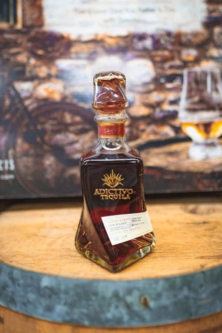 Adictivo 黑色陳年法國橡木桶釀製龍舌蘭酒 壁紙 — 一瓶Adictivo Tequila放在木桶頂部，旁邊有玻璃杯和質樸的石牆背景。