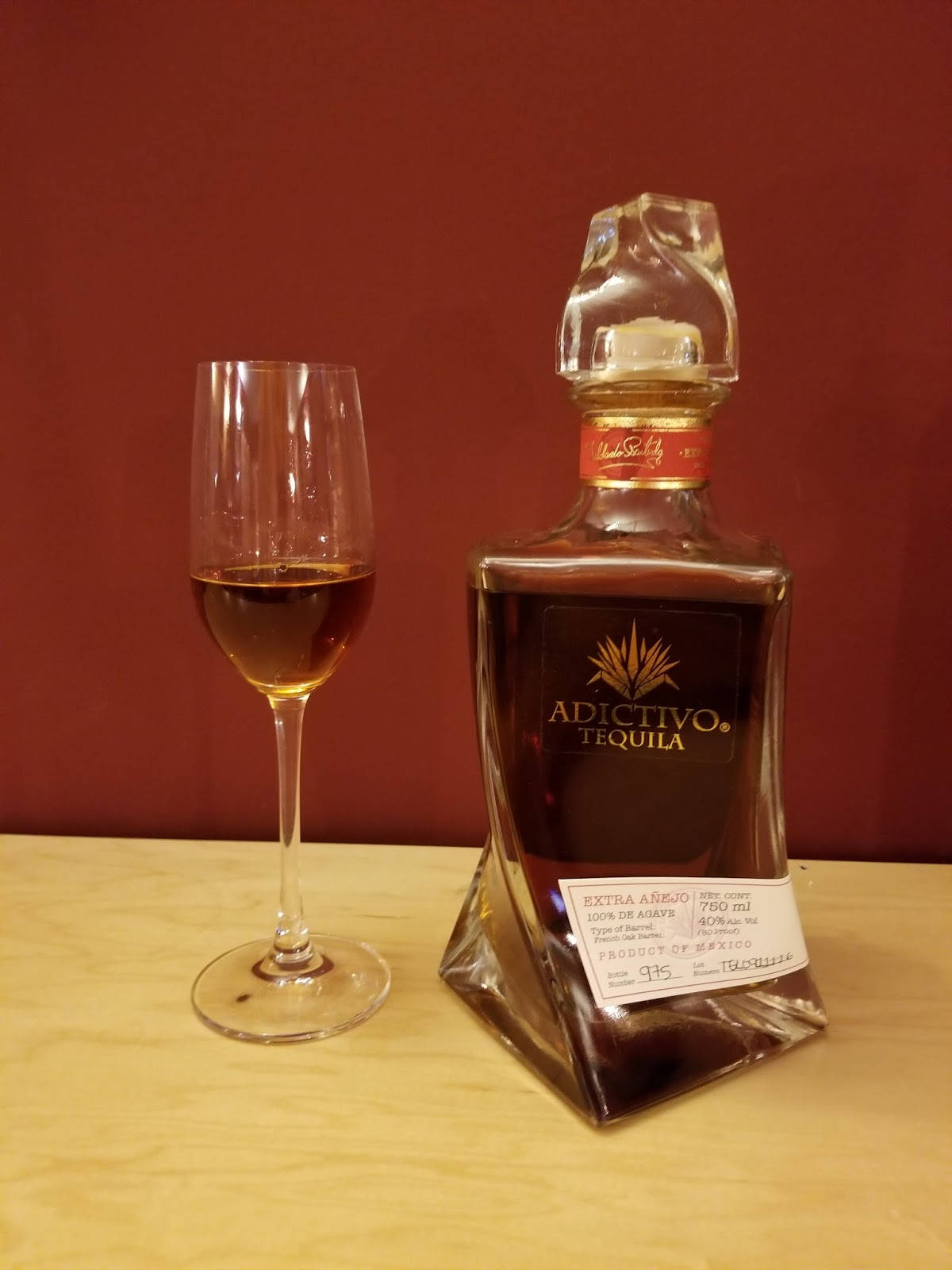 Adictivo極致黑桶陳釀龍舌蘭酒 壁紙 — 一瓶Adictivo Tequila Extra Añejo放在木質表面上，旁邊有一個裝滿酒的玻璃杯，背景是深紅色牆壁。