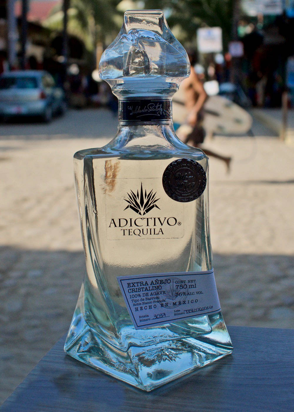 Adictivo水晶龍舌蘭酒 壁紙 — 一瓶透明的Adictivo Tequila玻璃瓶放在戶外的桌子上，背景是模糊的海濱街道場景。