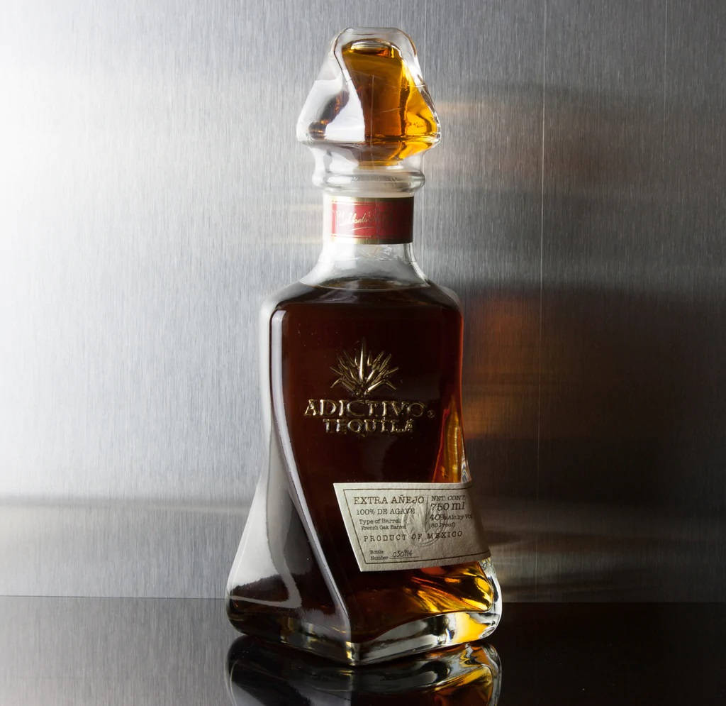 Adictivo白蘭地陶瓷龍舌蘭酒瓶 壁紙 — Adictivo Tequila Extra Añejo 的時尚玻璃瓶在拉絲金屬背景下閃耀著琥珀色液體。