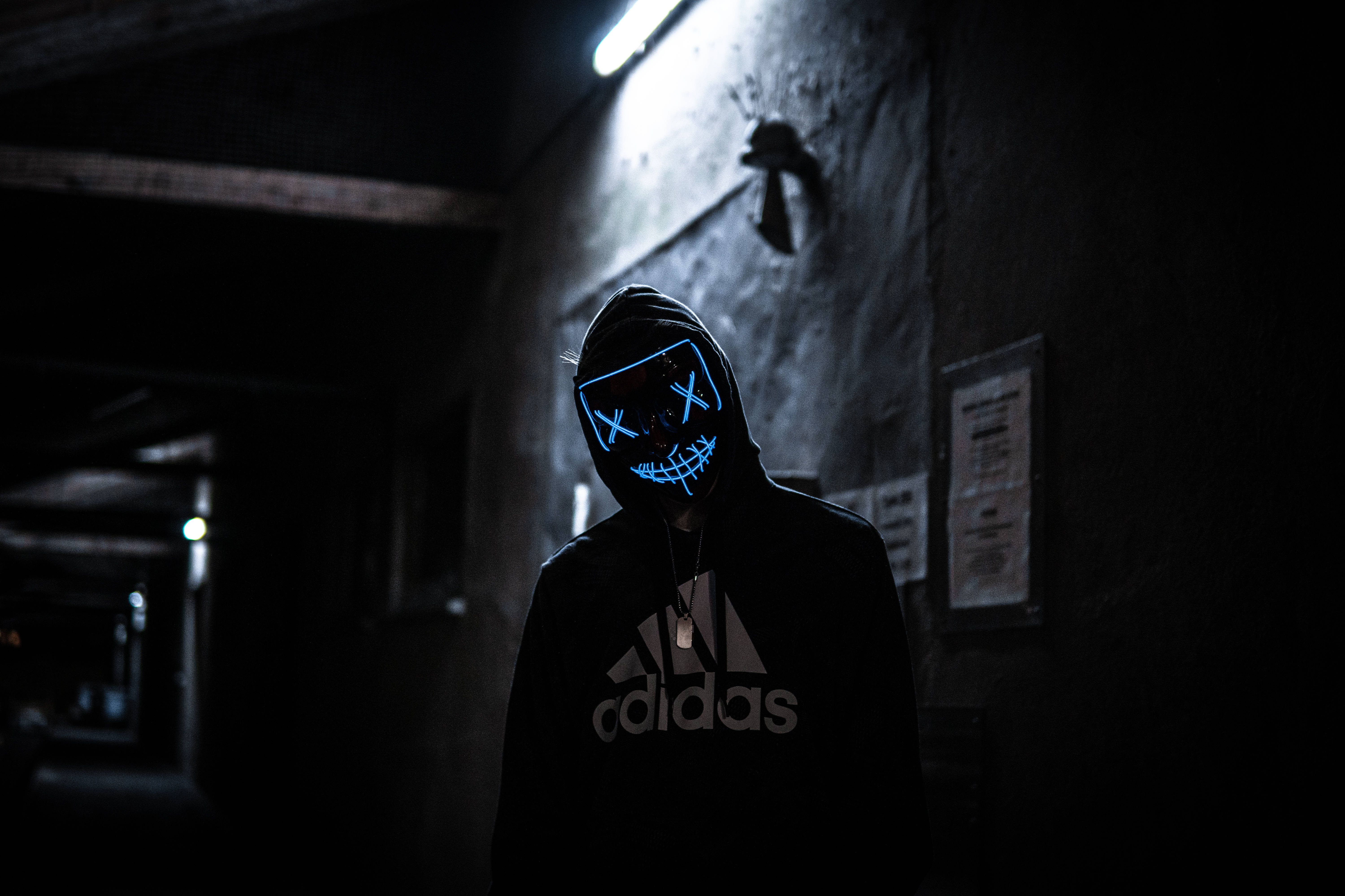 穿Adidas連帽衫的《清道夫》迷 壁紙 — 一個穿著黑色連帽衫的人戴著發光藍色LED面具，面具上有X形眼睛和咧嘴笑，站在光線昏暗的工業走廊中。