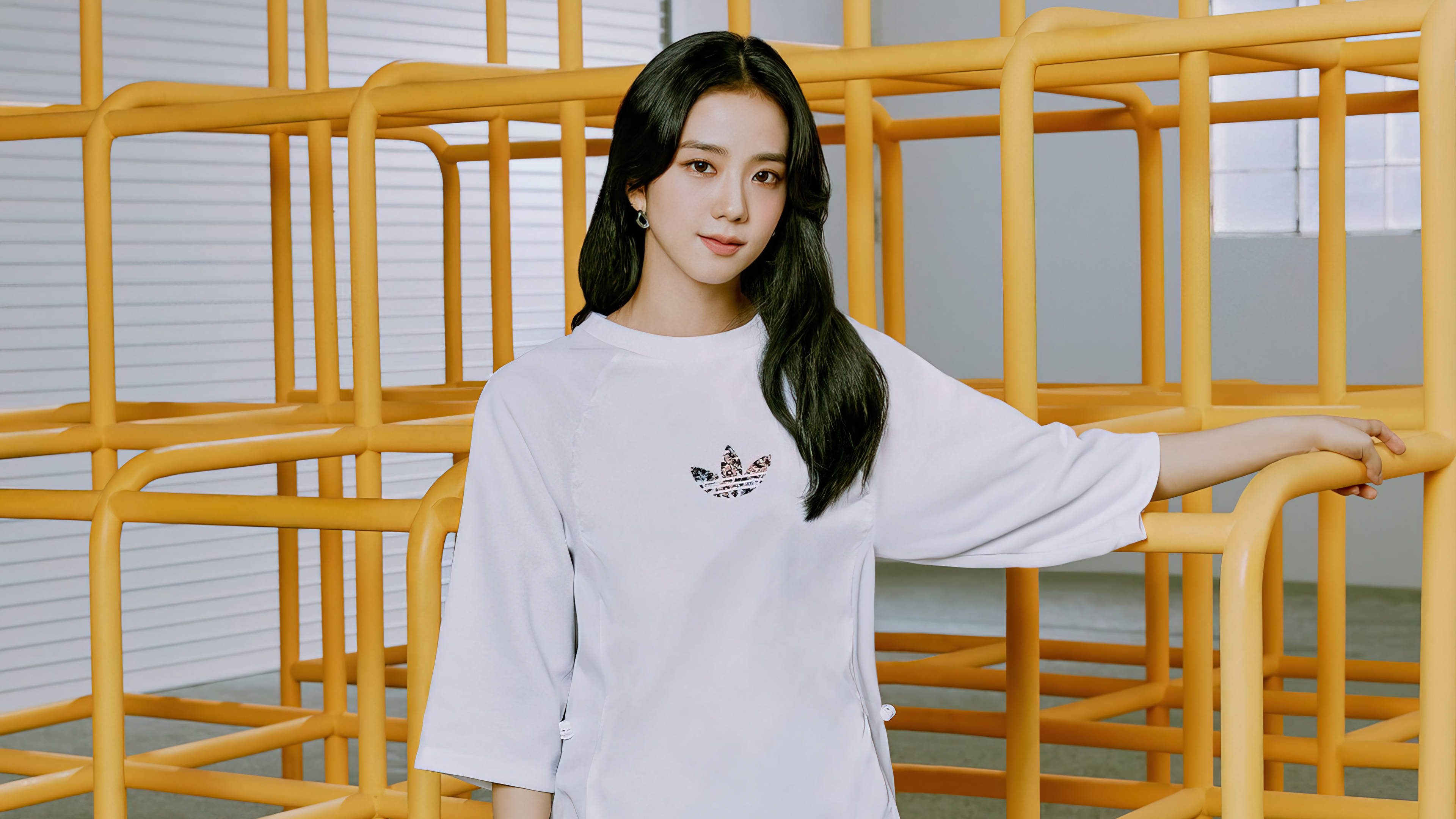 Adidas代言智秀 壁紙 — 一位長黑髮的年輕女子靠在亮黃色的金屬欄杆上，身穿帶有小圖案的白色T恤。