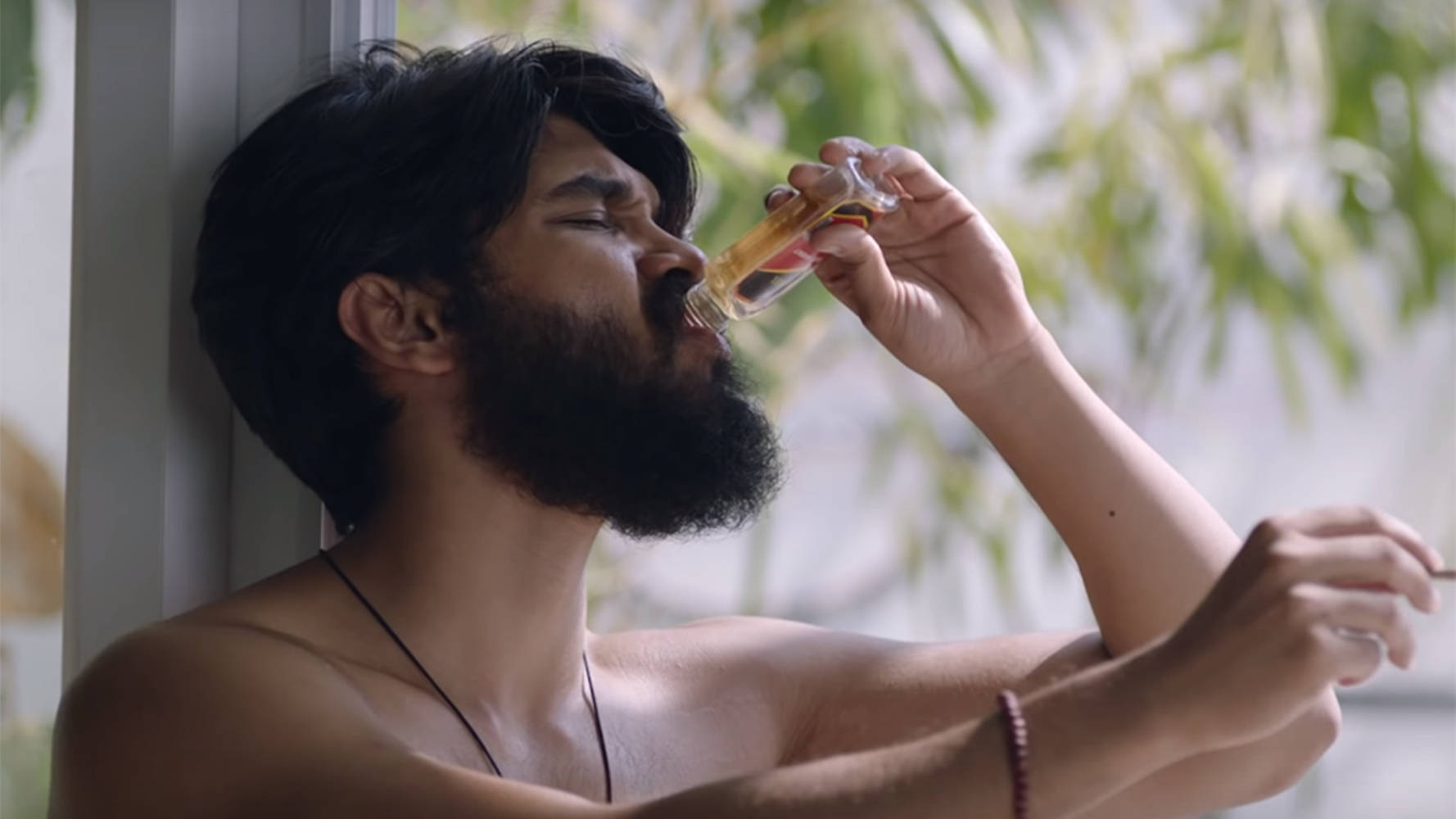 一個留鬍子的男人靠在窗邊，從玻璃瓶啜飲。
