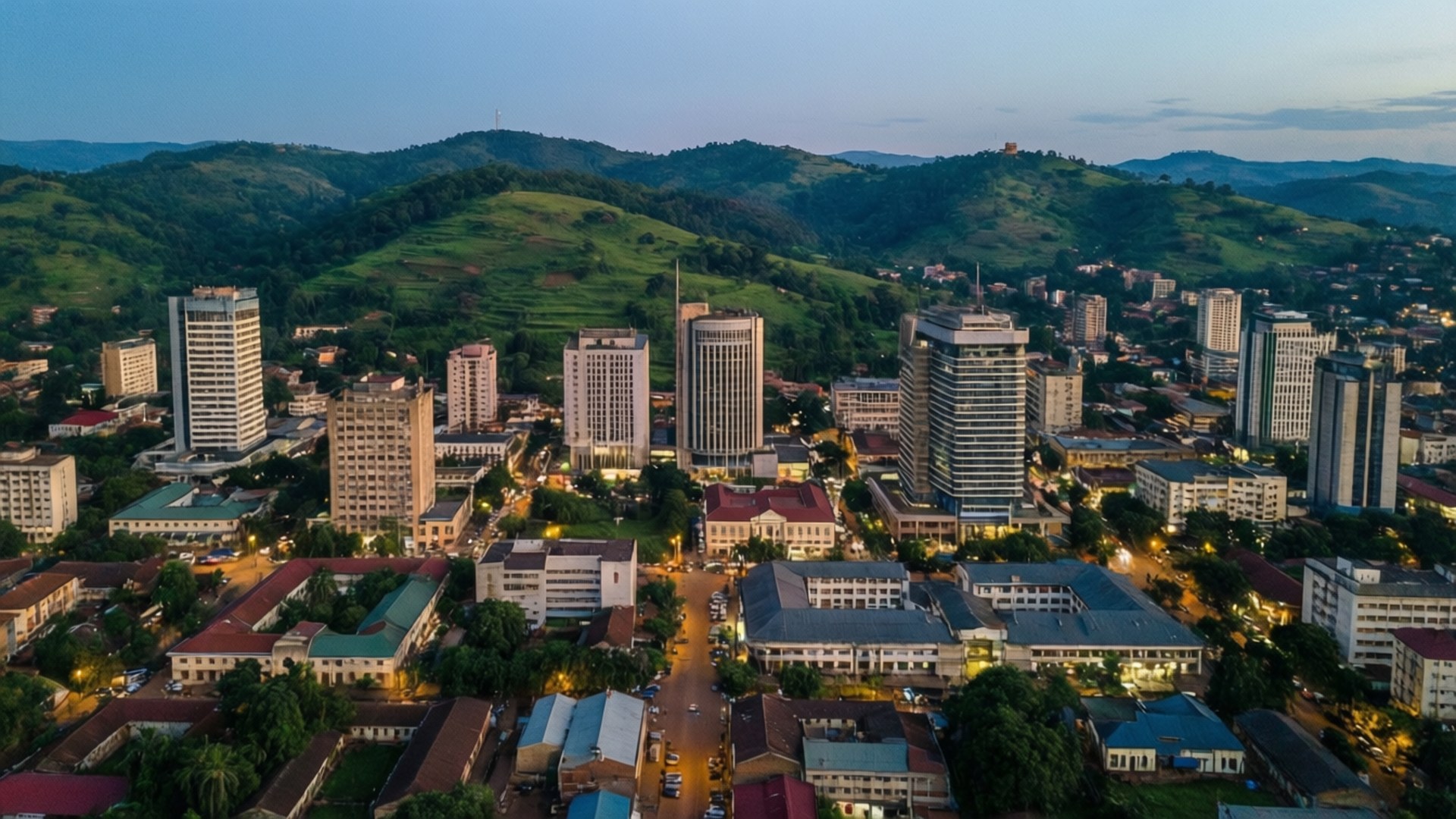 Yaoundé Skyline Evening 壁紙 — 鳥瞰一座坐落在鬱蔥青山之間的城市，暮色中街道燈火通明，現代建築林立。