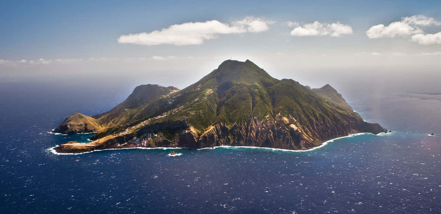 Caribbean Island 桌布 壁紙 — 在部分多雲的藍天下，一個崎嶇的綠色島嶼的鳥瞰圖，擁有陡峭的懸崖和碧綠的海水。