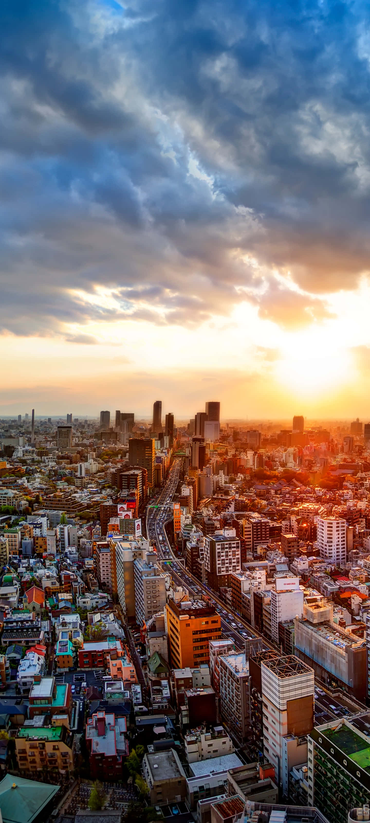 Panorama 桌布 壁紙 — 日落時分廣闊城市景觀的鳥瞰圖，金色光線穿過戲劇性的雲層，照射在密集的城市建築之上。