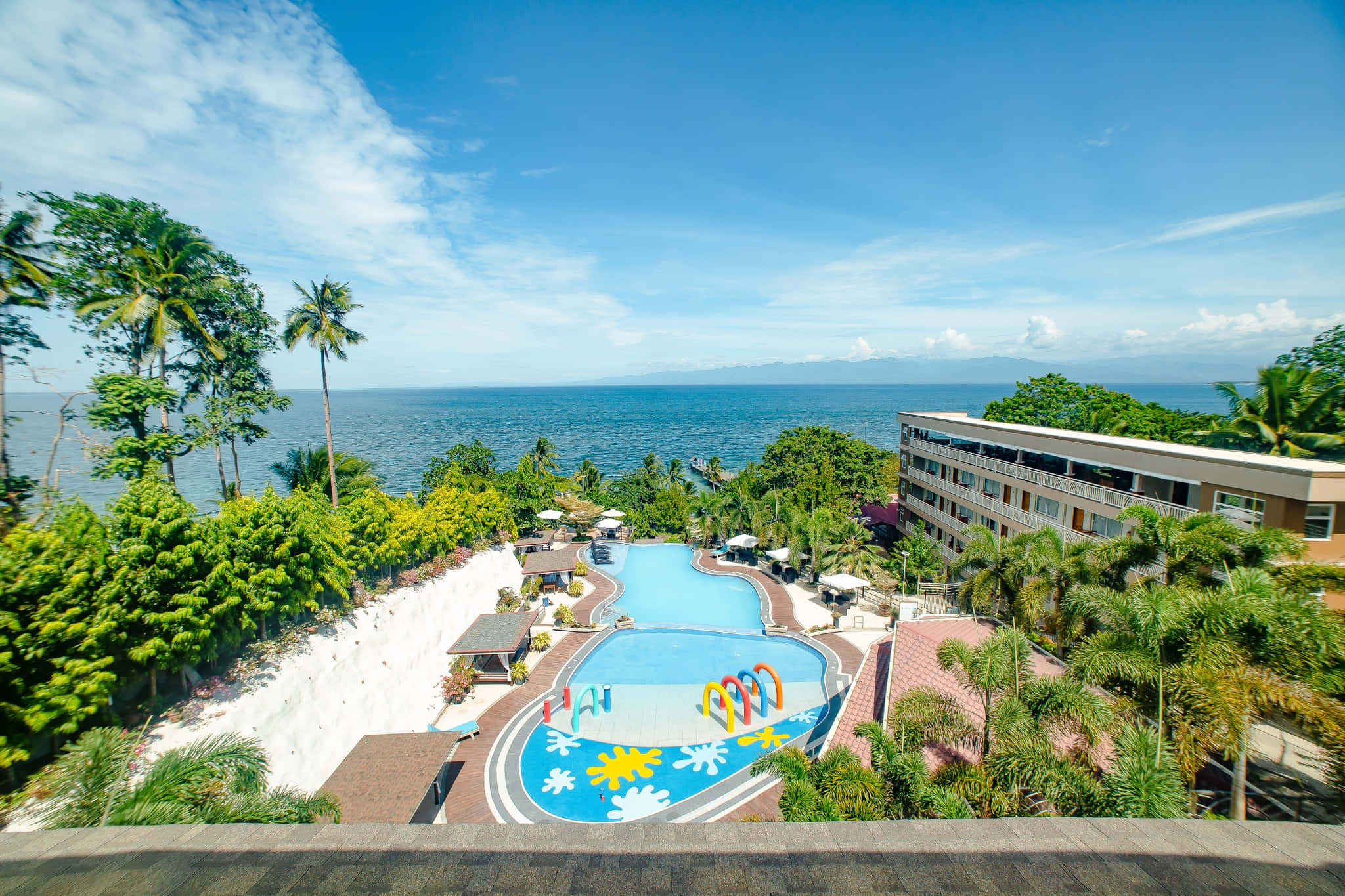 Beach Resort 桌布 壁紙 — 熱帶度假村的鳥瞰圖，蜿蜒的泳池和棕櫚樹俯瞰著海洋，天空湛藍。