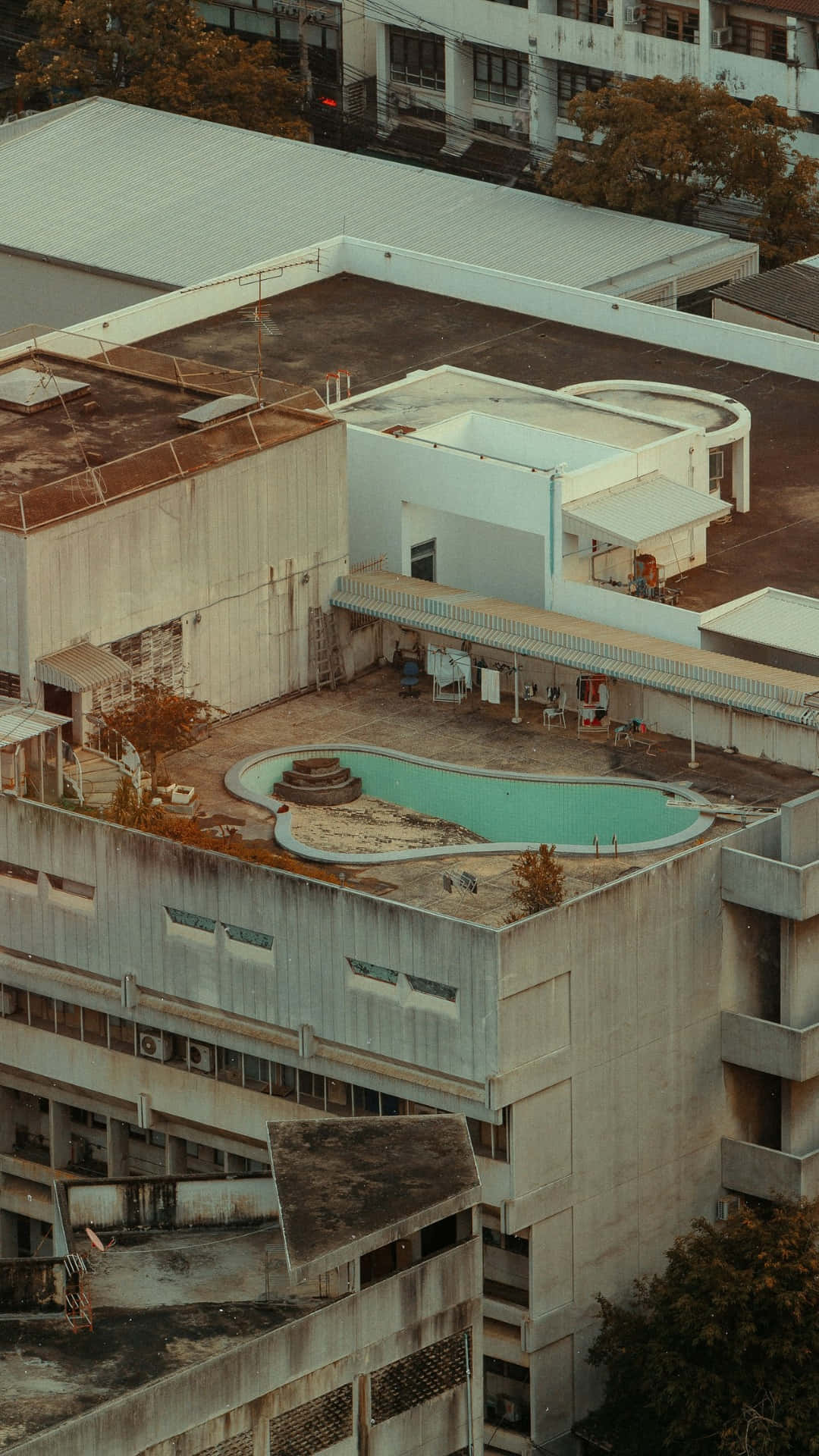  壁紙 — 從高空俯瞰，一個風化的天台泳池被混凝土建築物和秋天的樹木環繞，沐浴在溫暖的金色光線中。