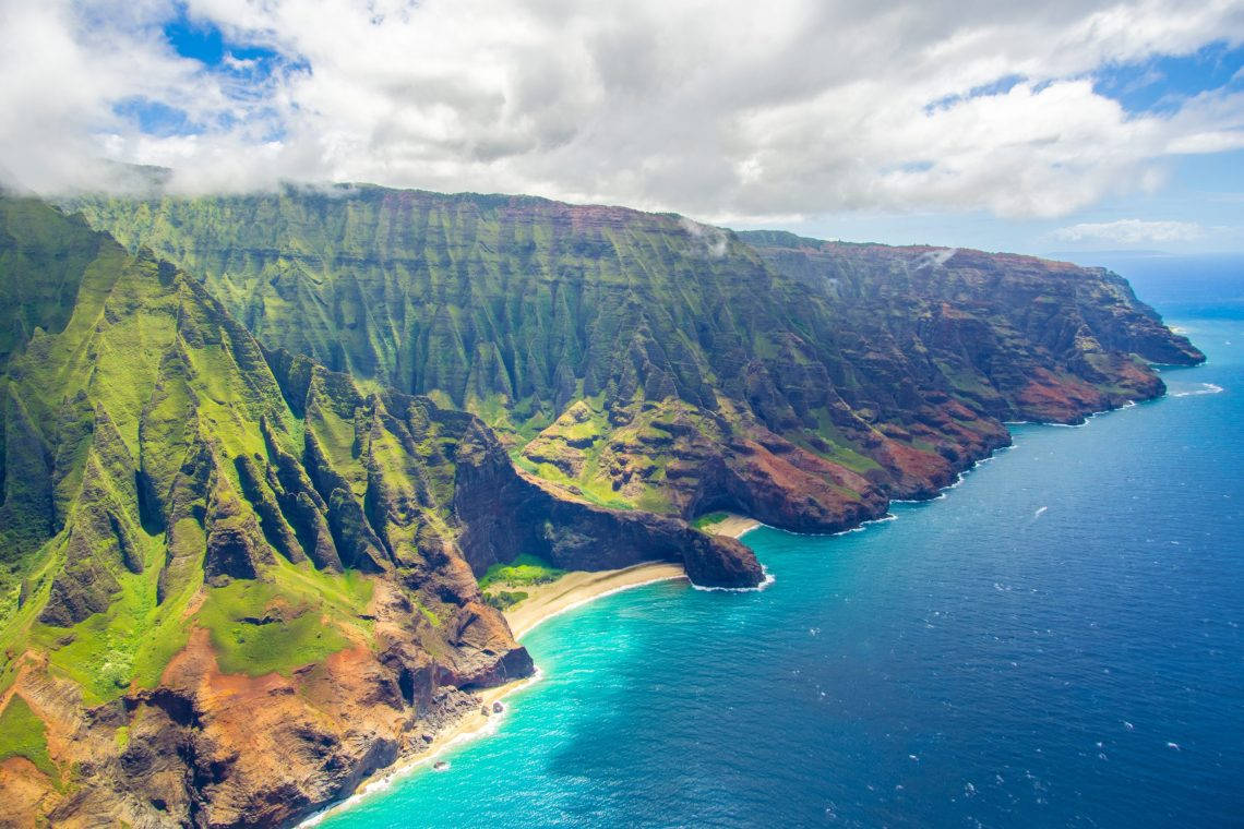 奧胡島多雲山脈 壁紙 — 戲劇性的綠色懸崖直插碧綠海洋，下方藏著一個沙質小海灣的航拍視圖。