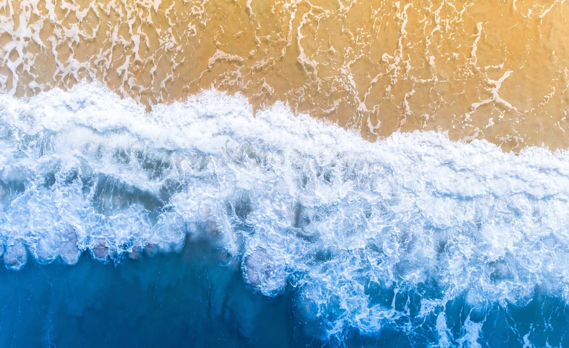  壁紙 — 從高空俯瞰海浪拍打金色沙灘，泛起白色泡沫和深藍色海水。
