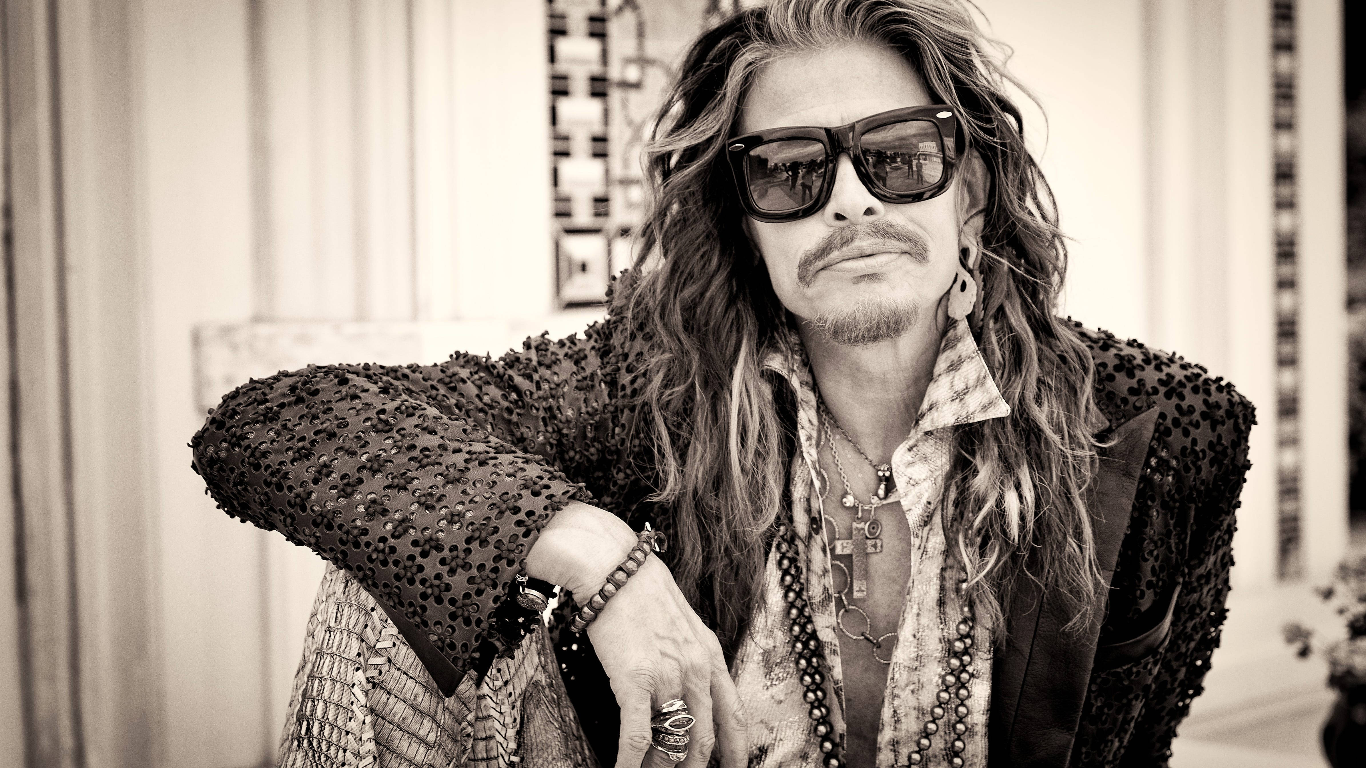 Aerosmith 樂隊史蒂芬·泰勒復古風 壁紙 — 一位長髮戴太陽眼鏡的時尚男士，穿著質感外套和層疊項鍊，自信地擺姿勢。