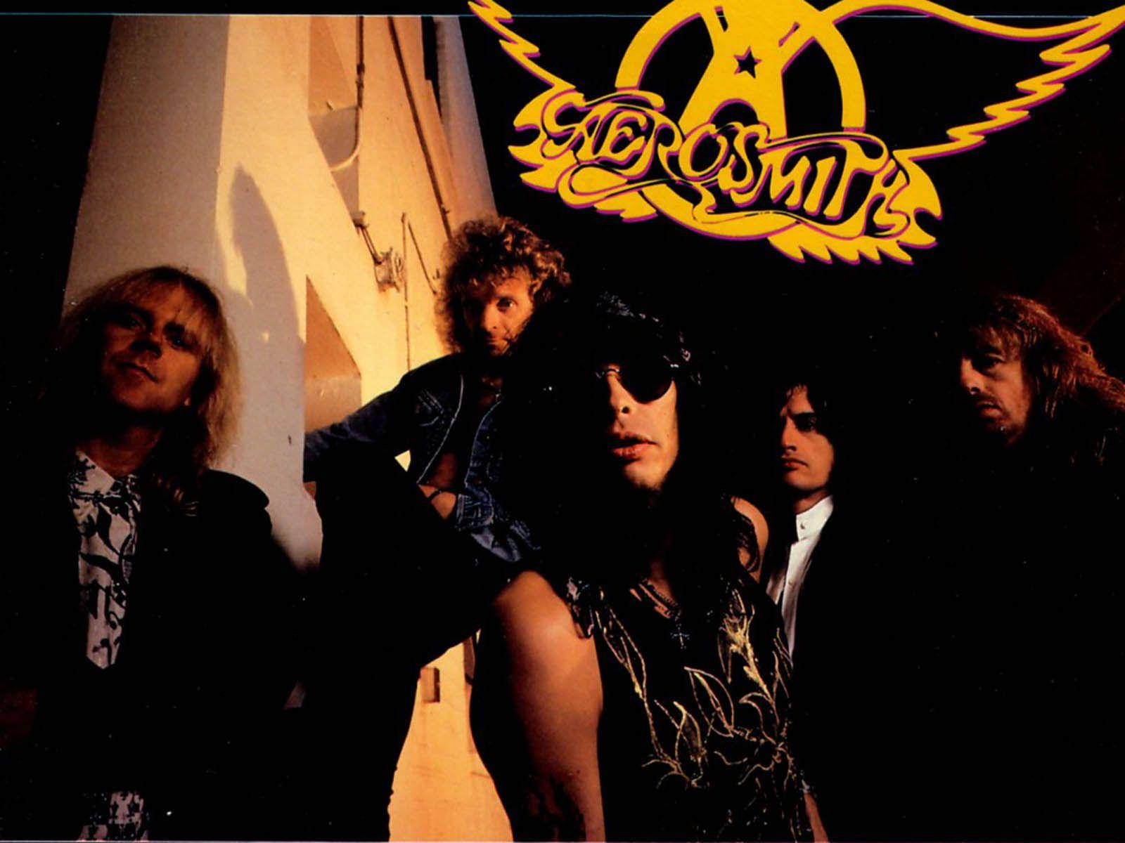 Aerosmith搖滾樂隊展現其深邃美學 壁紙 — Aerosmith 樂隊成員在溫暖燈光下戲劇性地擺姿勢，他們標誌性的翅膀標誌在上方發光。