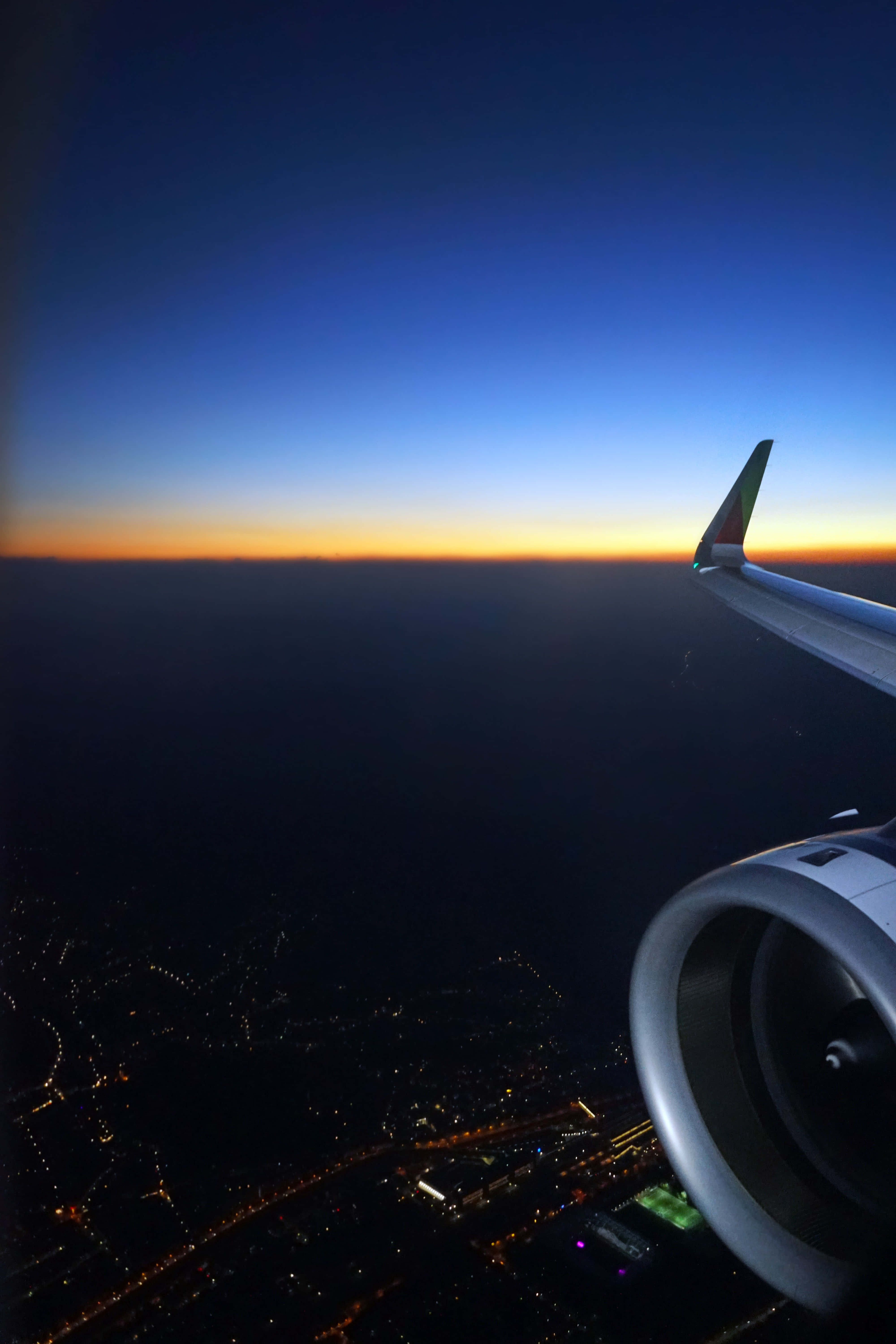 aerospace進展的壯闊一瞥 壁紙 — 飛機機翼和引擎在暮色天空和橙色地平線下的城市景觀中形成剪影。