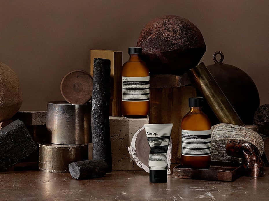 Aesop 美妝產品靜物 壁紙 — Aesop護膚產品展示在質樸的金屬和石頭物品之間，呈現溫暖的大地色調。