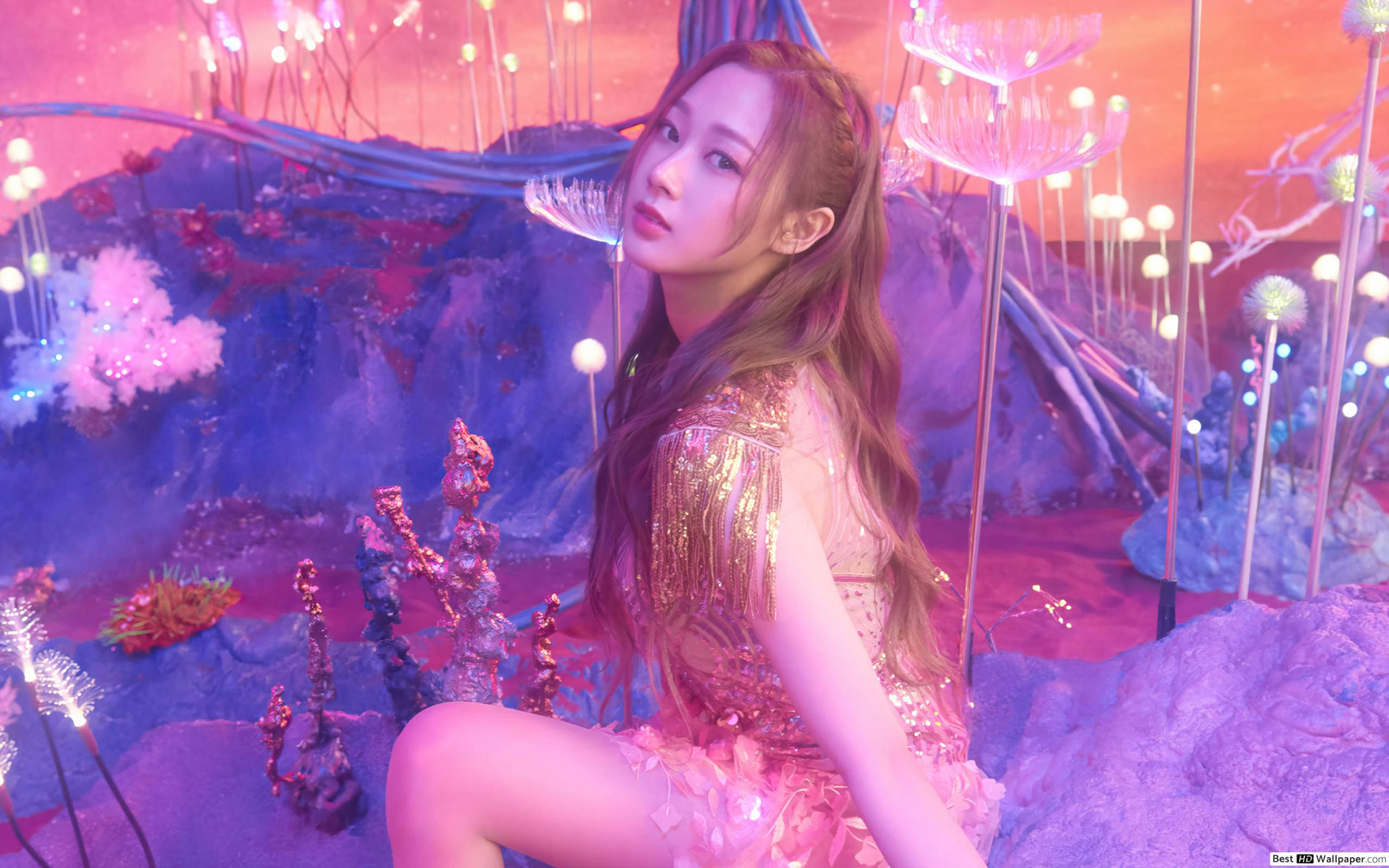 Aespa Winter 舞台震撼演出 壁紙 — 一位身穿閃爍金色服裝的女性優雅地坐在超現實的粉紅色和紫色外星風景中，周圍有發光的植物。