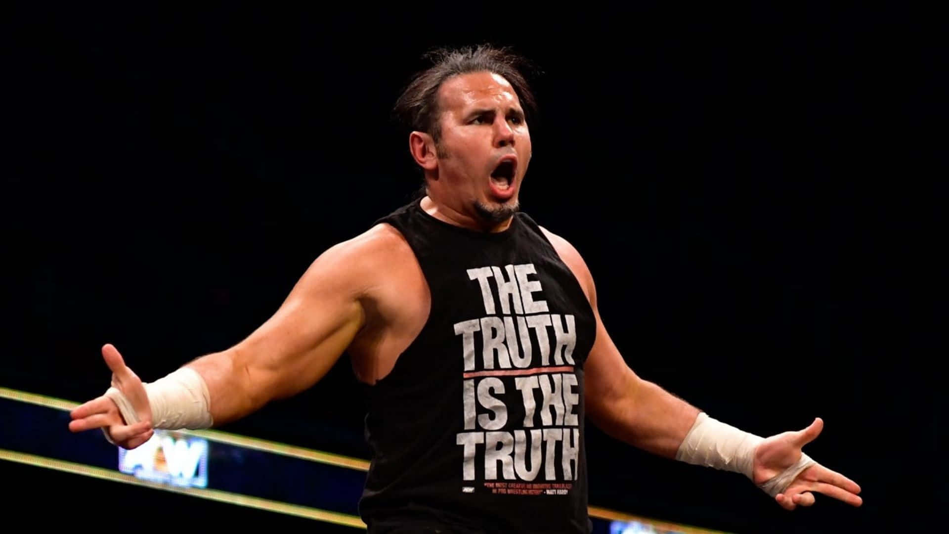 AEW 美國摔角手馬特·哈迪指槍動作 壁紙 — 身穿印有「THE TRUTH IS THE TRUTH」字樣的黑色背心的摔角手在明亮燈光下於擂台上張開雙臂咆哮。