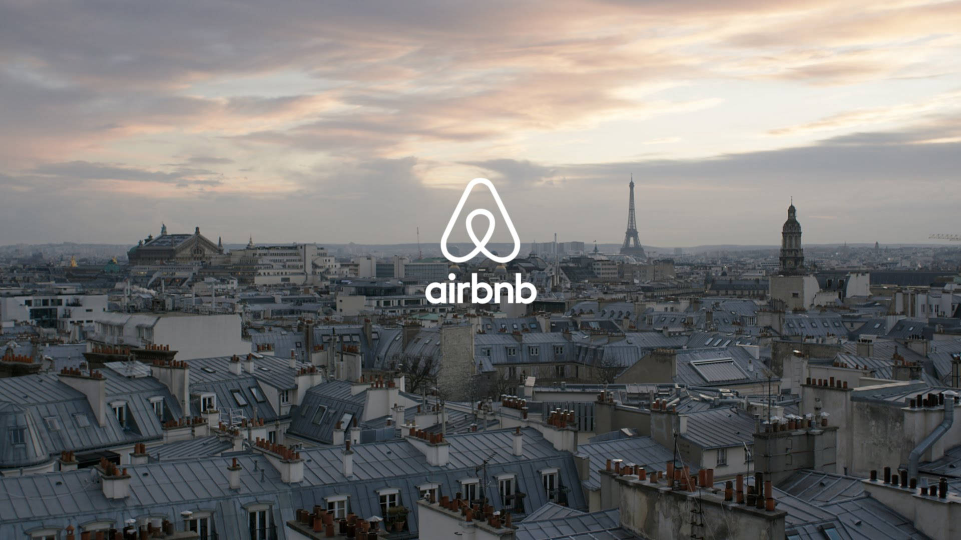 巴黎風景中的Airbnb標誌 壁紙 — 巴黎屋頂在柔和暮色下延伸，艾菲爾鐵塔清晰可見，Airbnb 標誌居中。