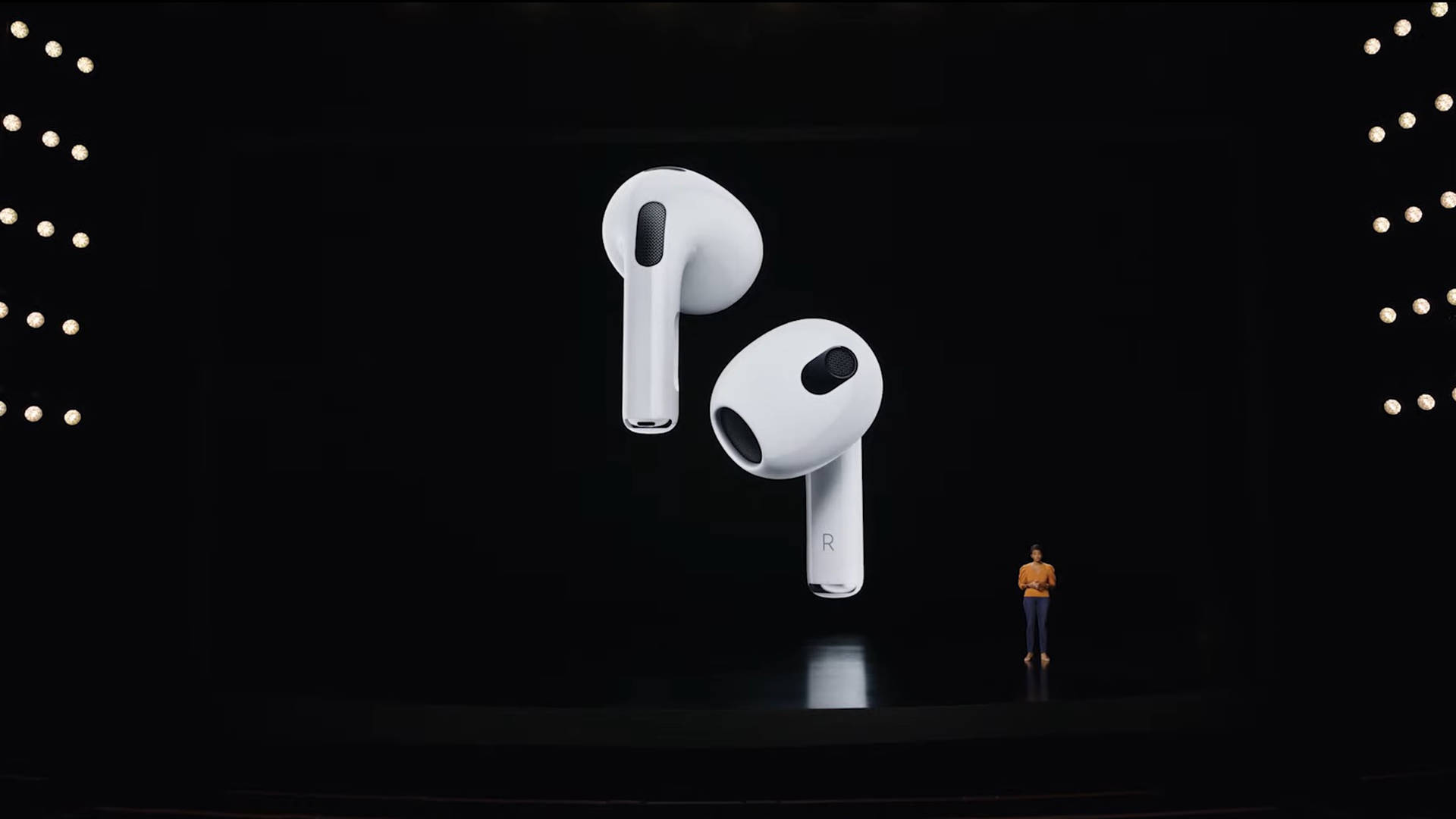 AirPods 第三代 壁紙 — 一位講者站在黑暗的舞台上，面前是一個巨大的屏幕，顯示兩側帶有發光燈光的浮動白色無線耳塞。