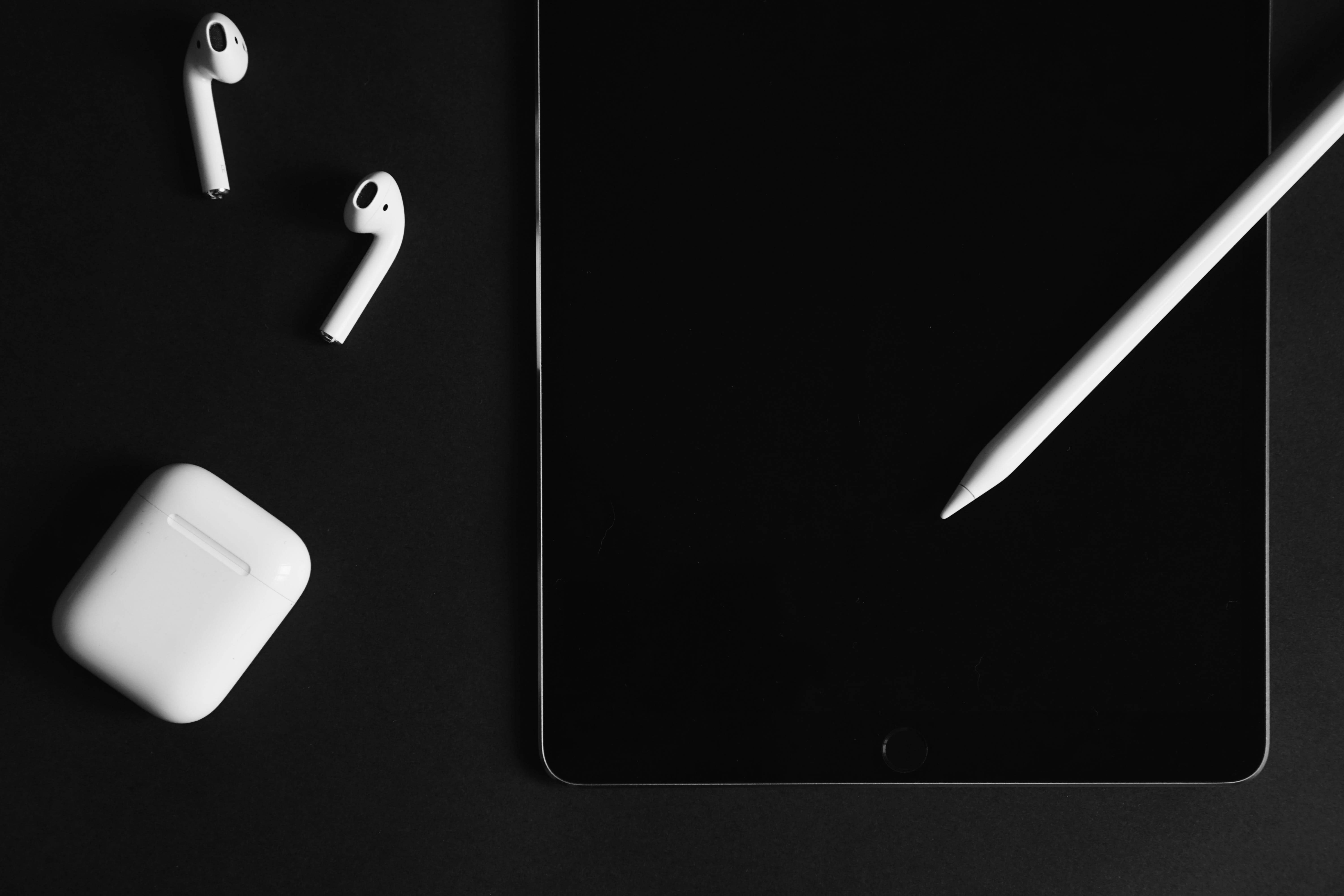 AirPods 與 iPad 壁紙 — 白色耳機盒和觸控筆放在時尚的黑色平板電腦上，背景是極簡的深色表面。