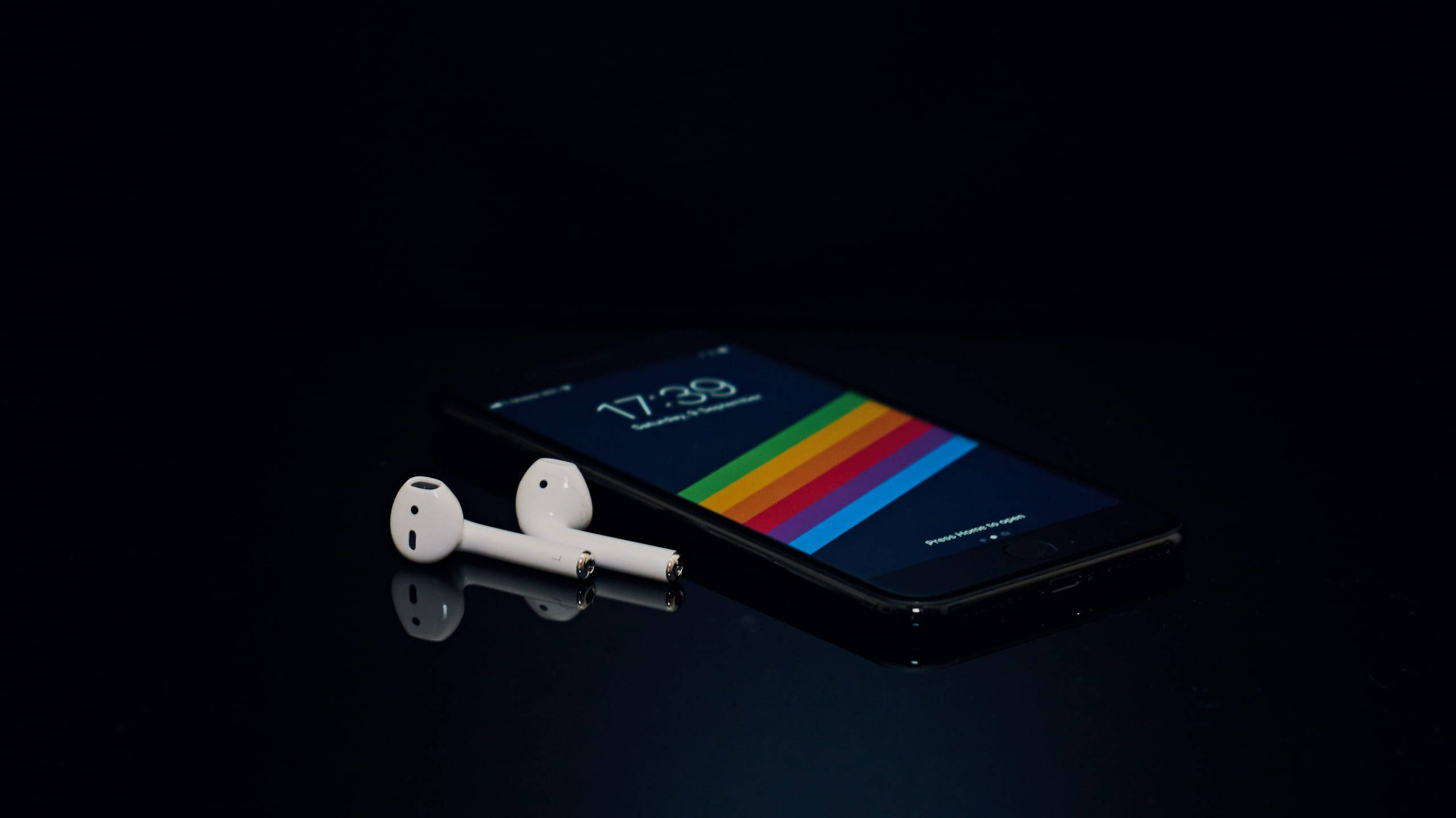 AirPods 與黑色 iPhone 7 壁紙 — 一部時尚的黑色智能手機，帶有彩虹條紋牆紙和白色耳塞，放置在反光的深色表面上。