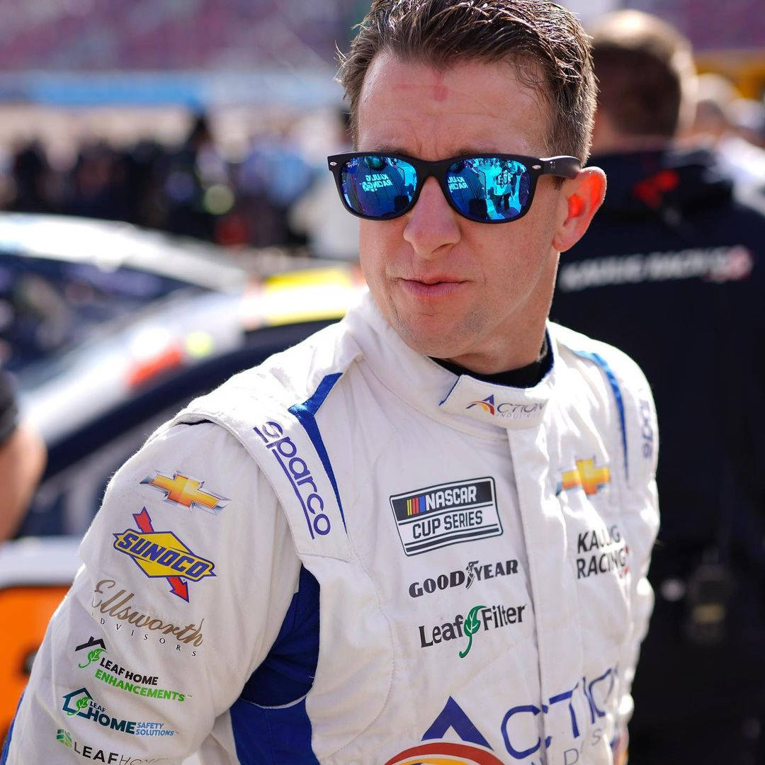 AJ Allmendinger 轉身回望 壁紙 — 一名NASCAR車手身穿白色賽車服和藍色太陽眼鏡，自信地站在賽車場上，背景是模糊的汽車和人群。