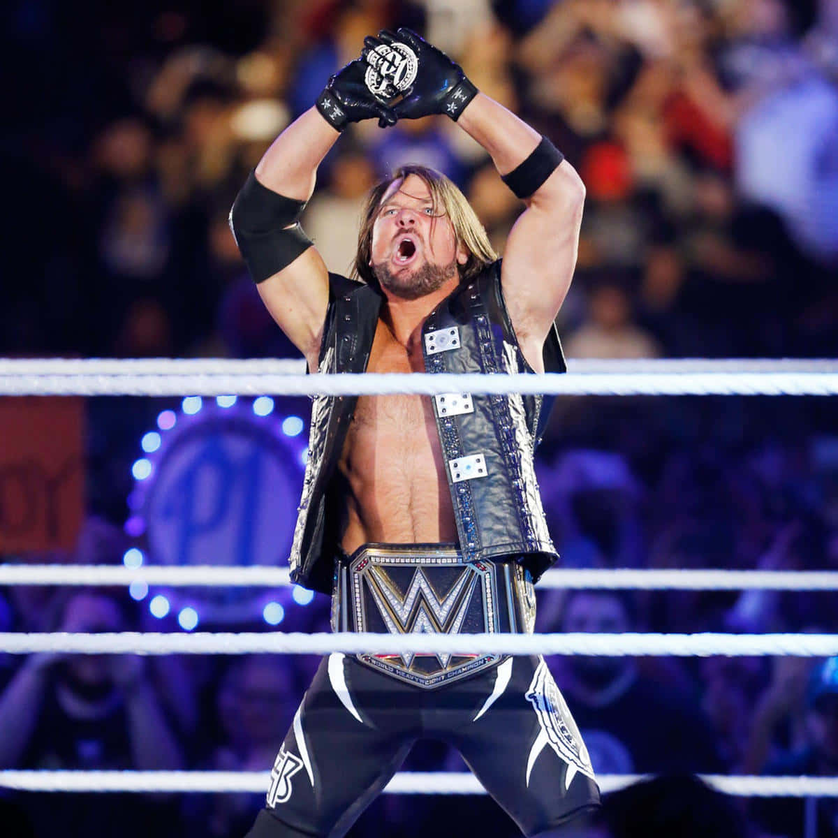 AJ Styles，The Club 領袖 壁紙 — 一位肌肉發達的摔角手穿著黑色背心和冠軍腰帶，在燈光明亮的摔角場內舉起戴手套的拳頭慶祝勝利。