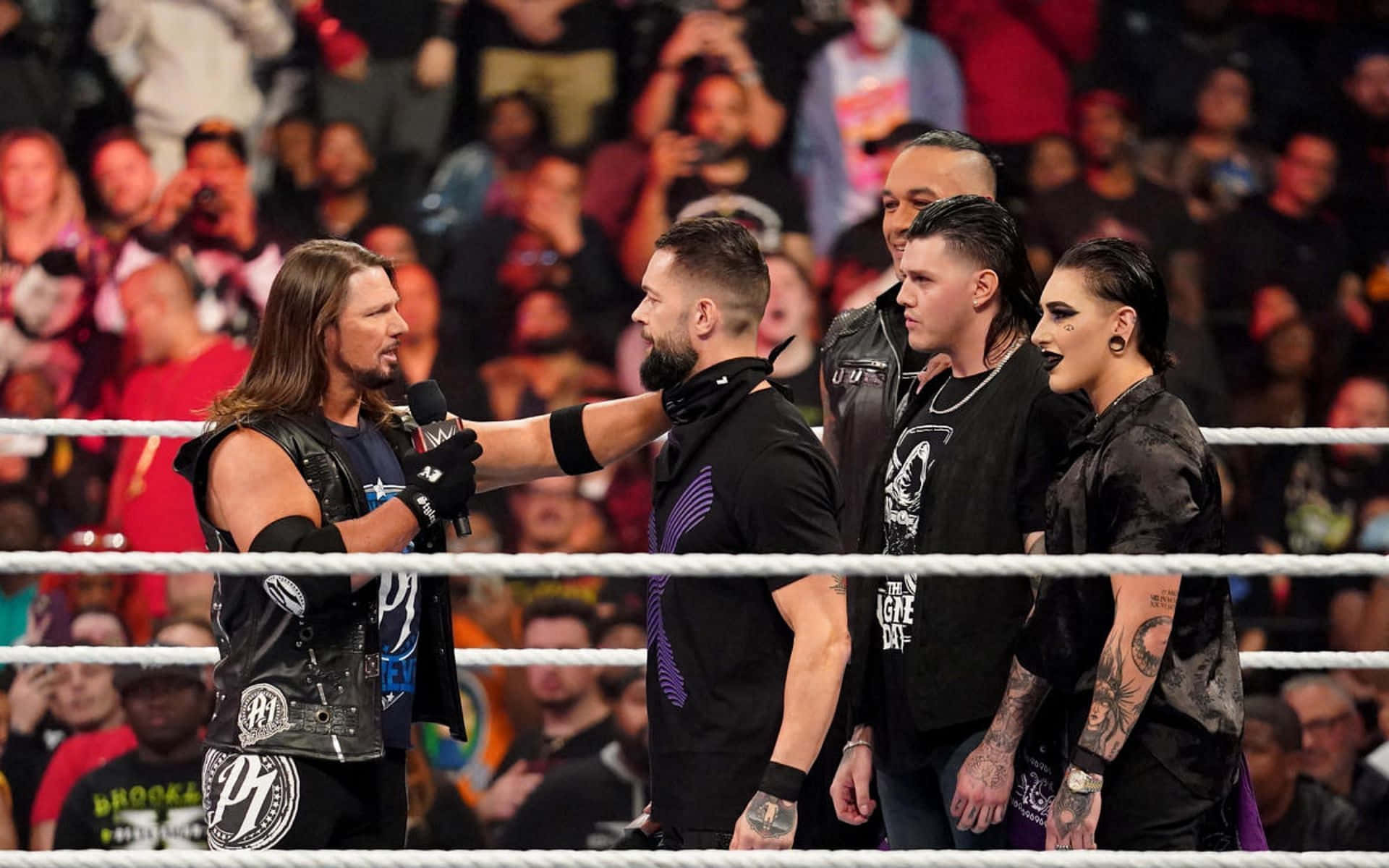 AJ Styles WWE Raw現場 壁紙 — 長髮摔角手拿著麥克風，在擂台燈光下與對手對峙，觀眾熱切觀看。
