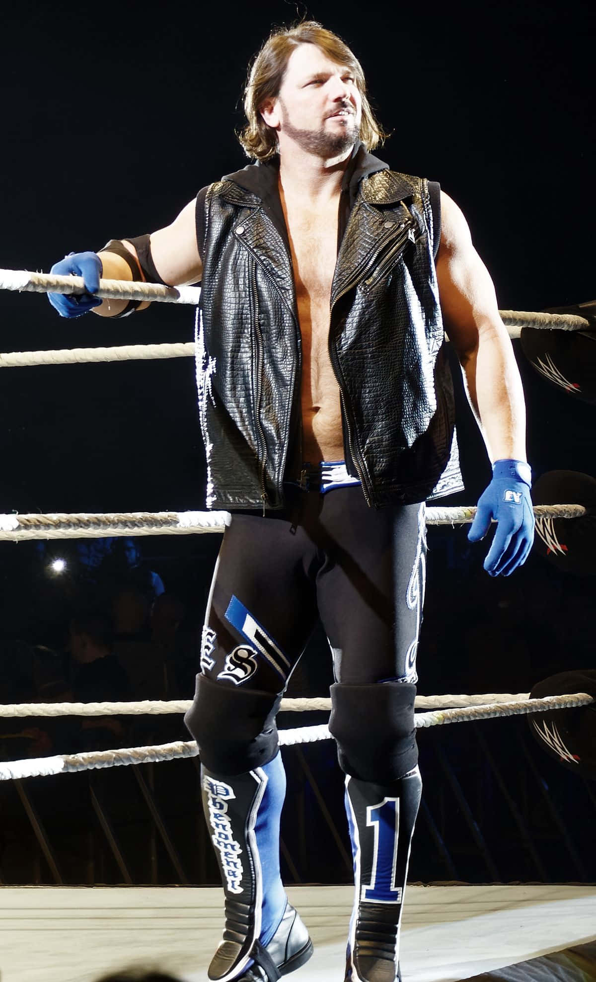 AJ Styles 世界重量級冠軍 壁紙 — 一位肌肉發達的摔角手，身穿黑色皮背心和藍色手套，在明亮的場館燈光下自信地站在摔角擂台內。