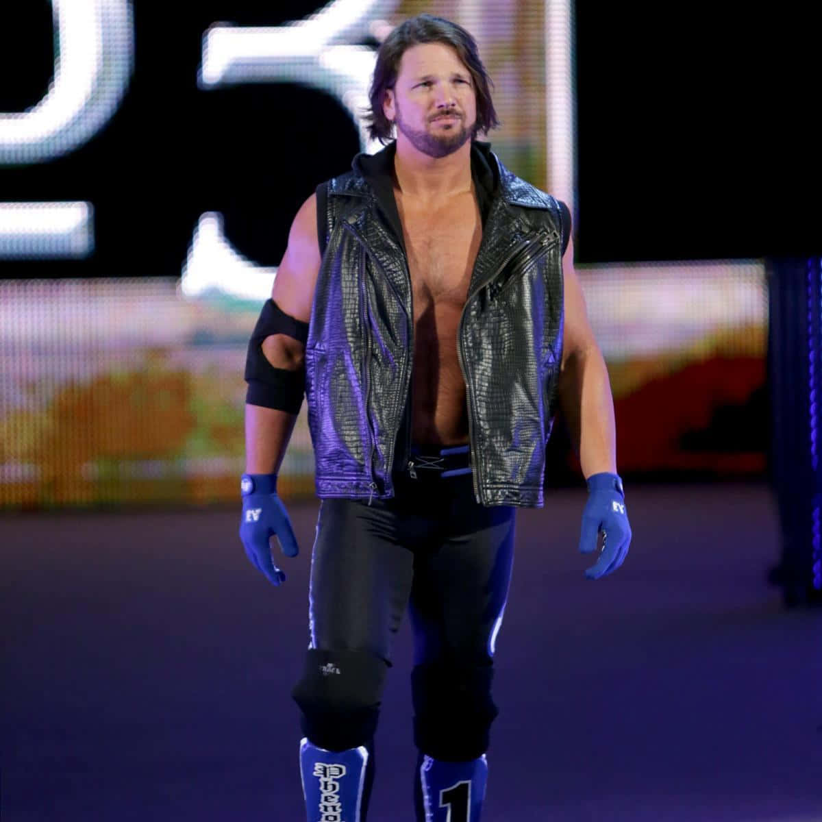 AJ Styles 亦稱「孤狼」 壁紙 — 一位肌肉發達的摔角手，身穿黑色皮背心和藍色手套，自信地走過燈光昏暗的競技場舞台。